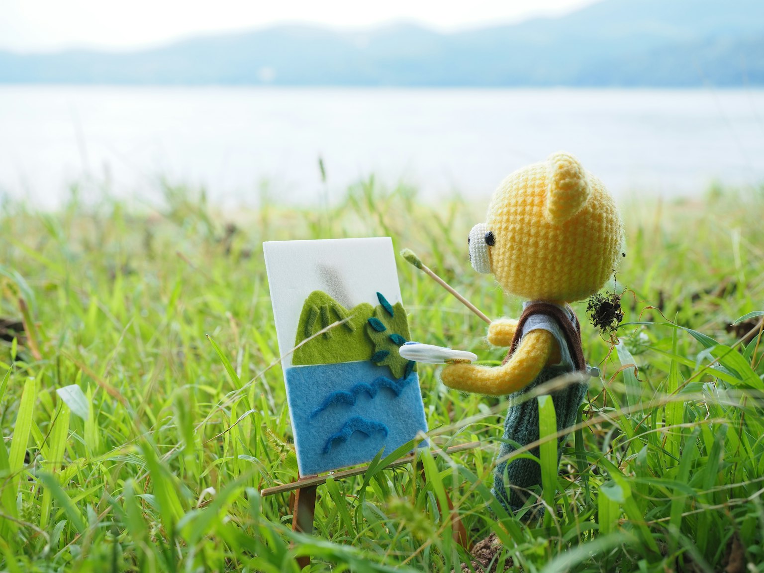 Un personnage en peluche d'ours peignant sur une toile dans l'herbe près d'un lac