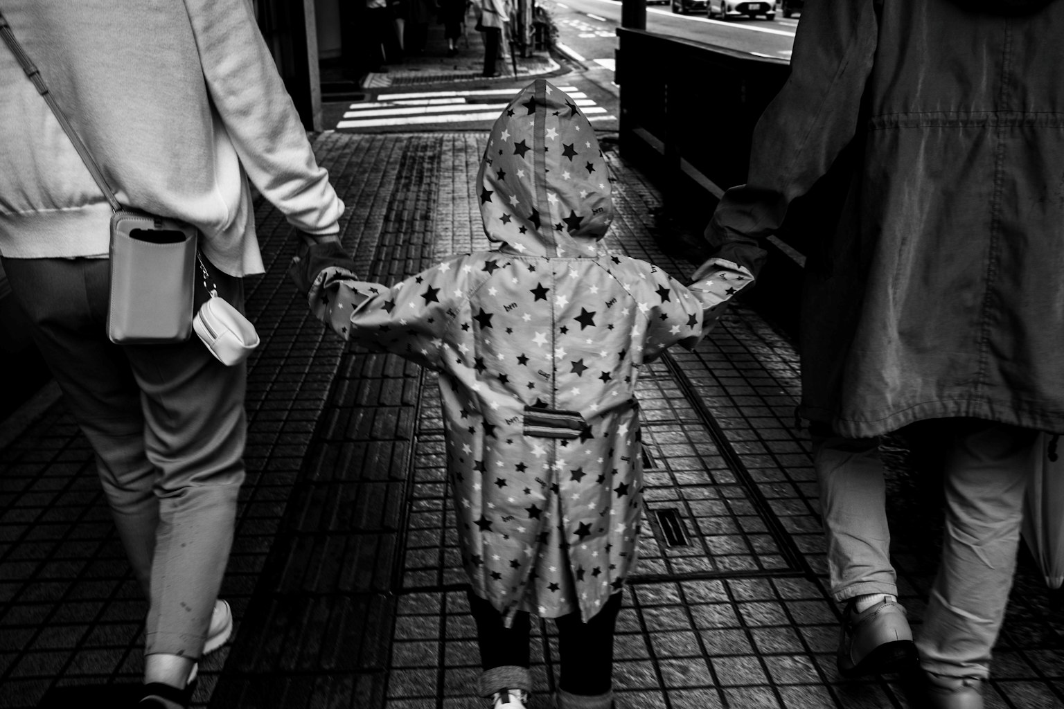 Foto hitam putih anak berpakaian jas hujan bergandeng tangan dengan orang tua saat berjalan