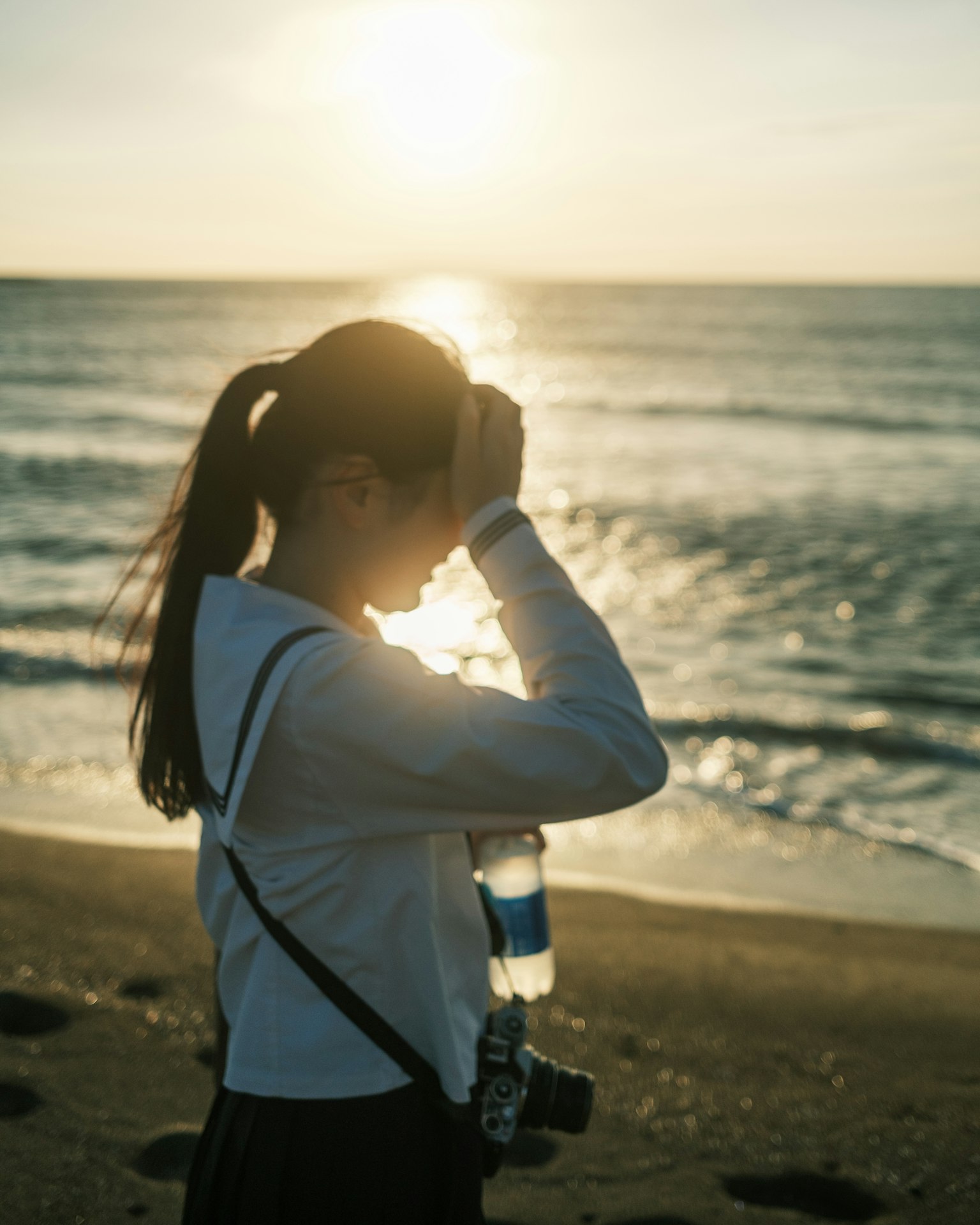Silhouette di una donna sulla spiaggia al tramonto che tiene una bottiglia d'acqua con una macchina fotografica a tracolla
