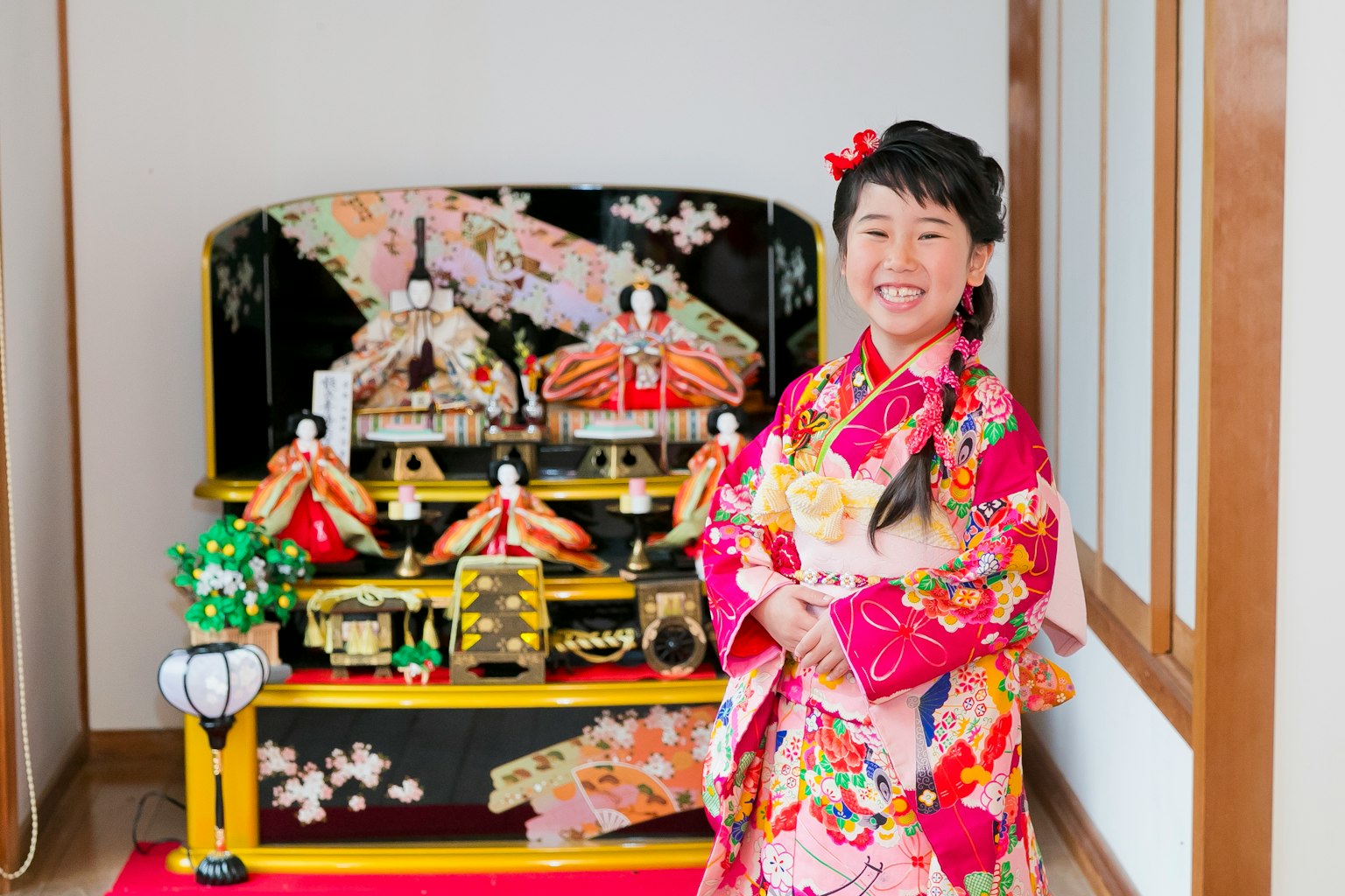 一个穿着色彩鲜艳和服的女孩微笑着，背景是传统的雏人形展示