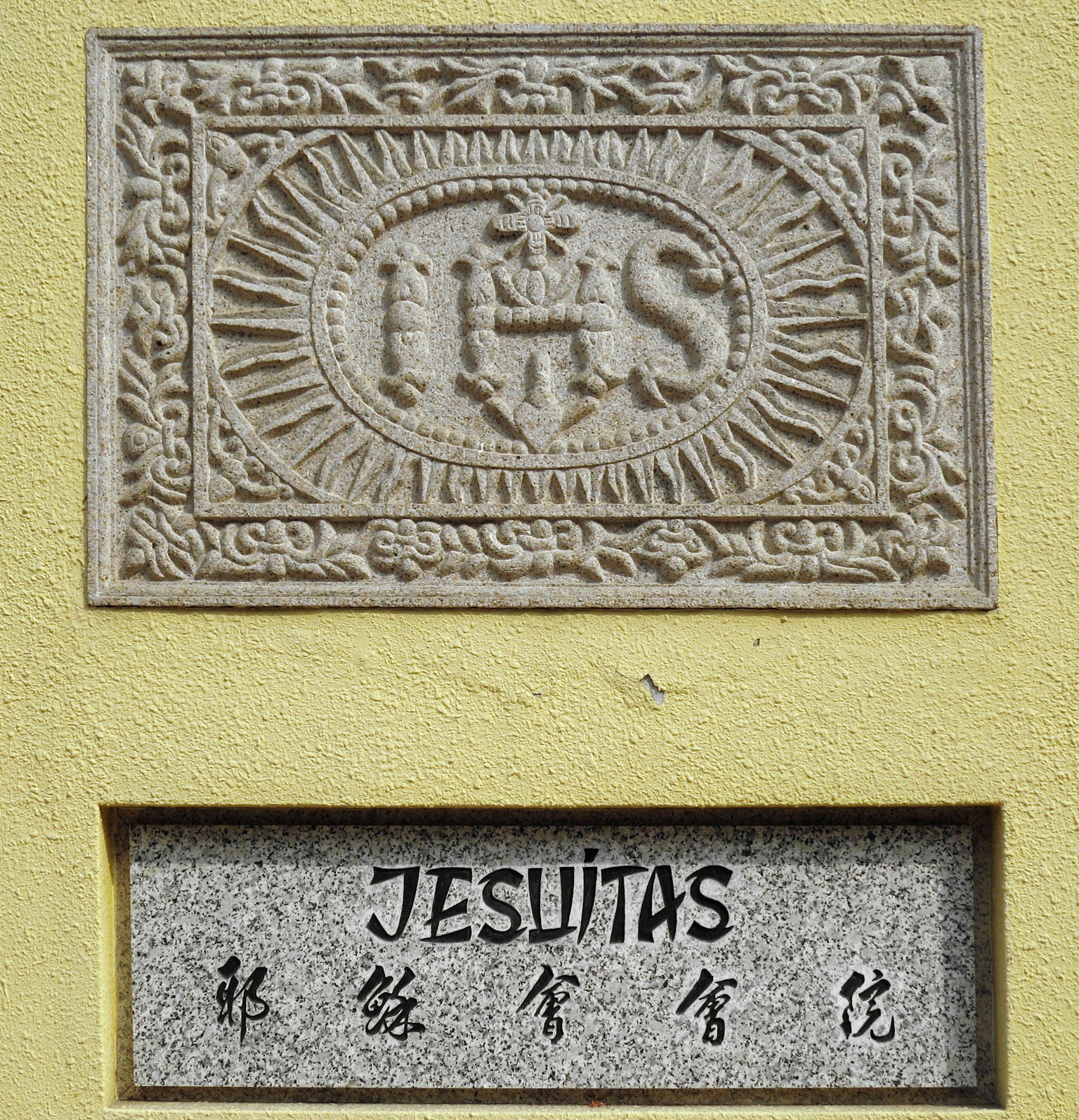 Emblema IHS scolpito e iscrizione JESUITAS su un muro giallo