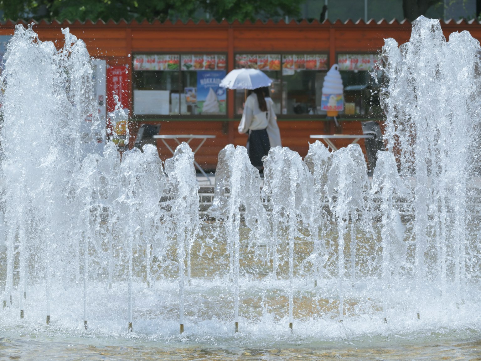 Eine Frau steht vor einem Wasserbrunnen mit einem Regenschirm und einem Essensstand im Hintergrund