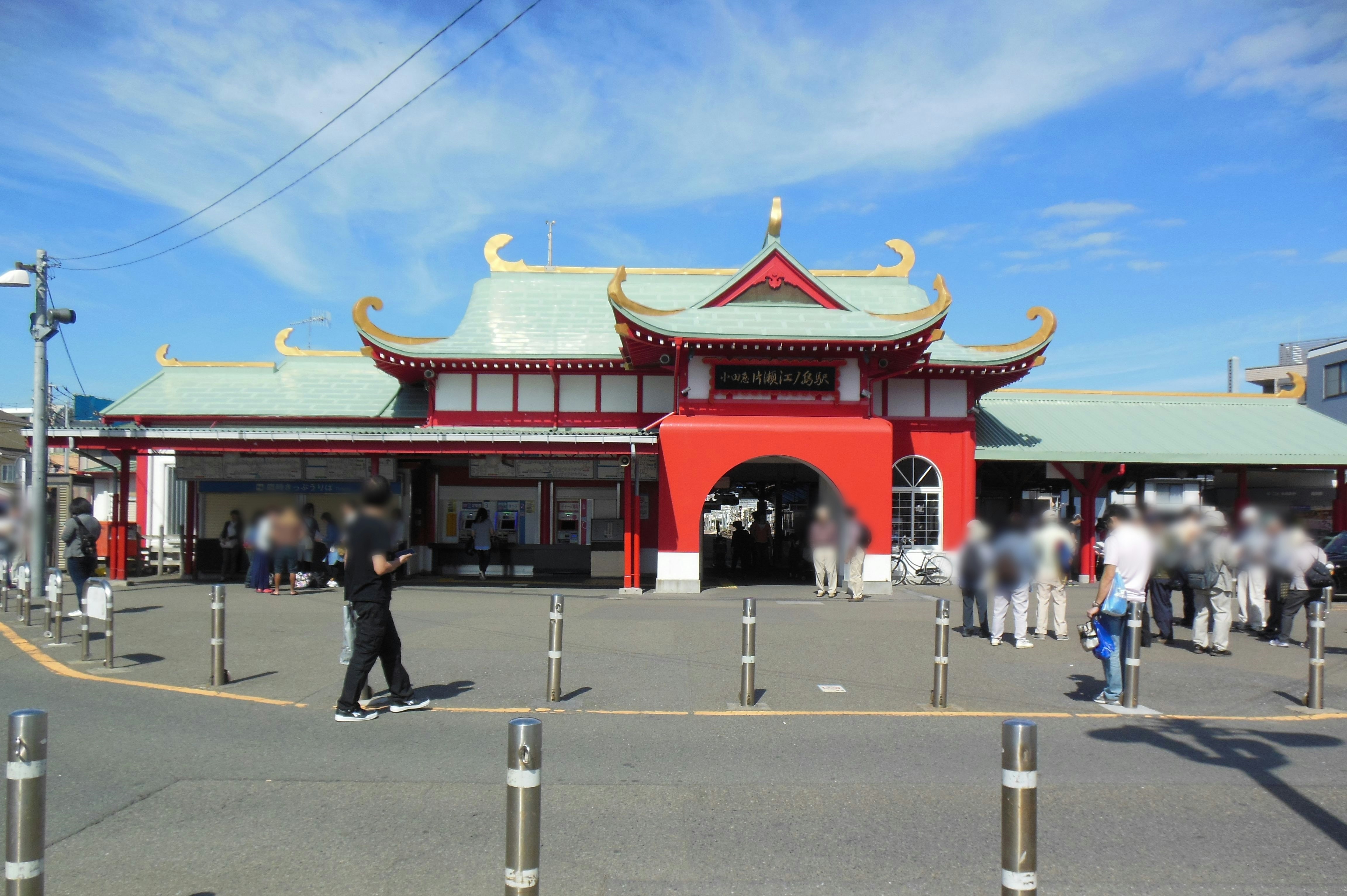 赤い門と屋根のある神社風の建物前で人が行き交う駅前の通り