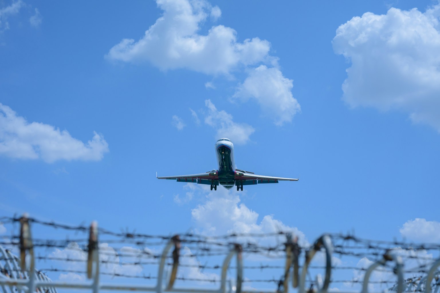 Immagine di un aereo in fase di atterraggio sotto un cielo blu con nuvole e filo spinato