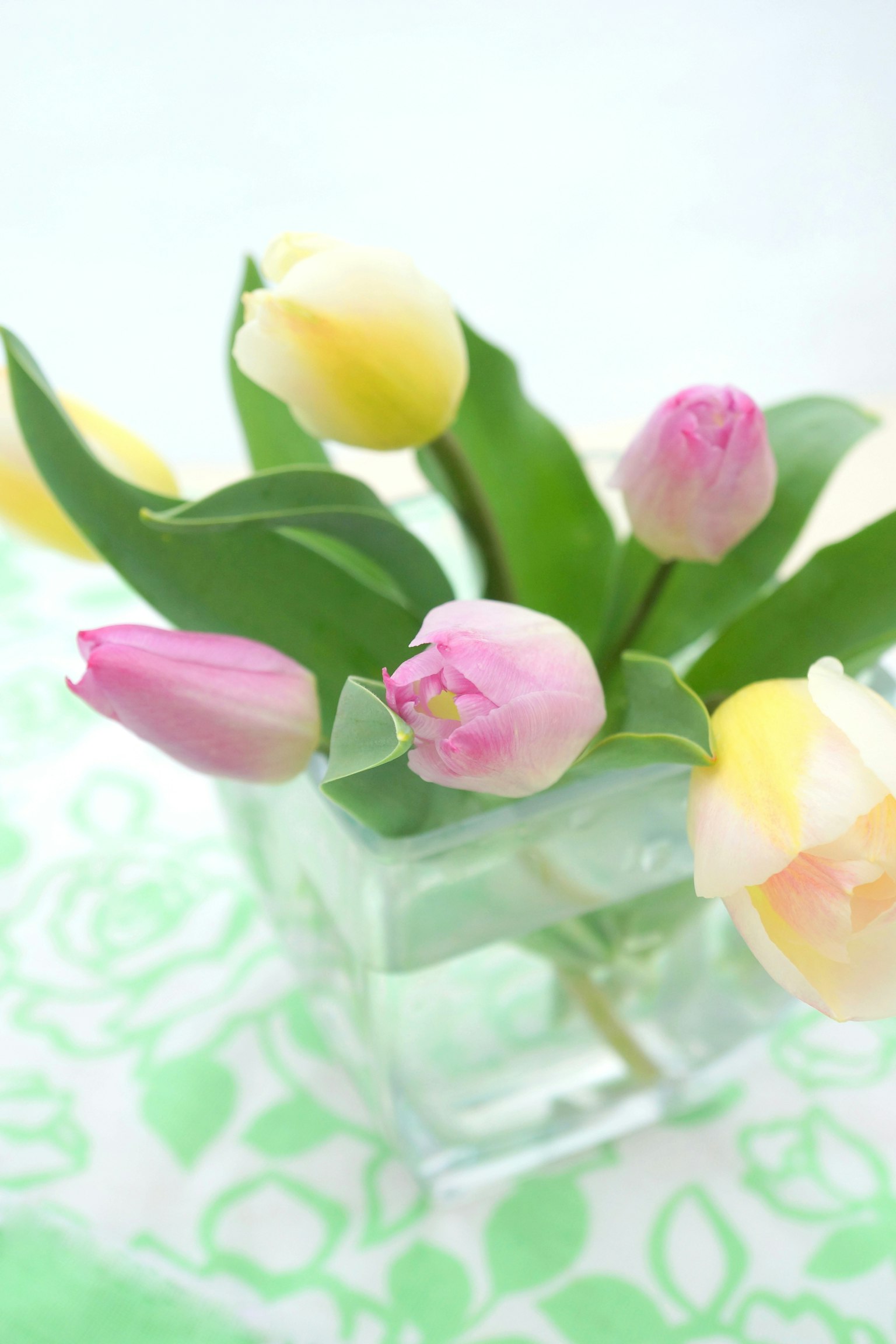 Un ramo de tulipanes rosas y amarillos en un jarrón transparente con hojas verdes y un fondo estampado