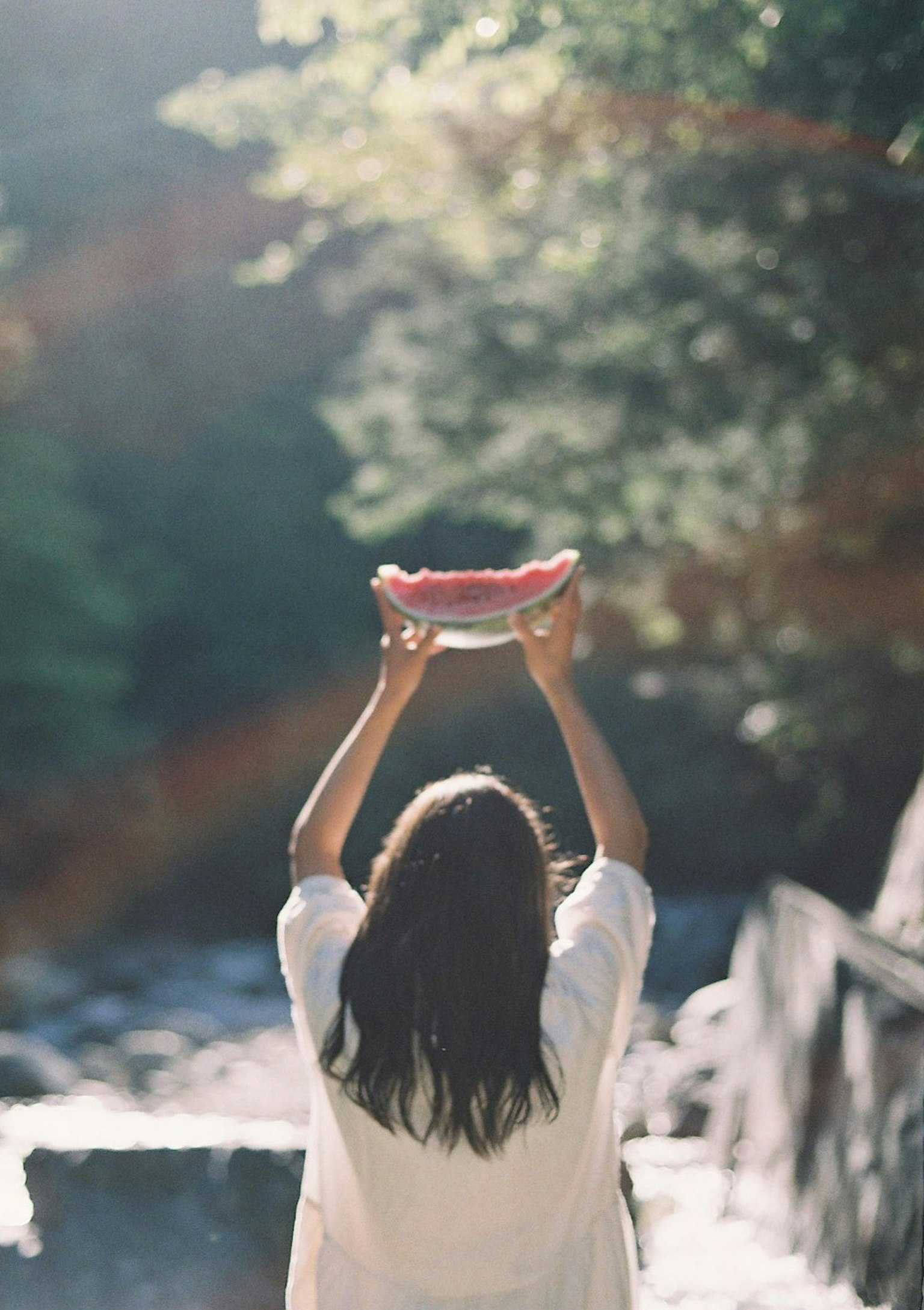 A woman holding a watermelon above her head in a natural setting with sunlight filtering through trees