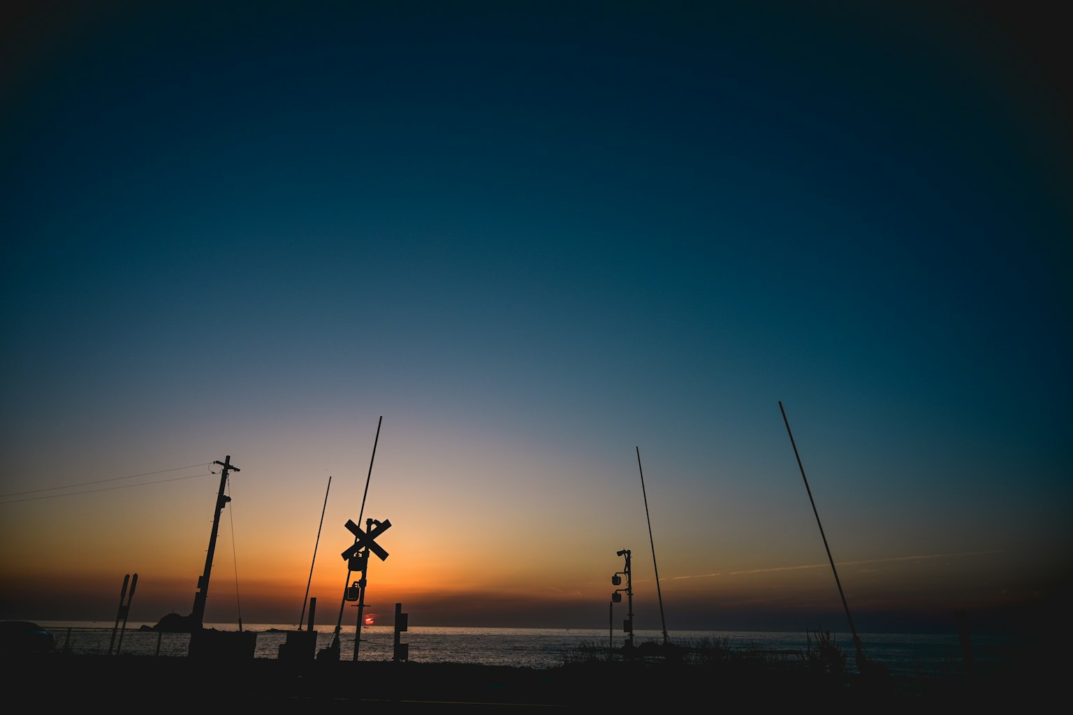 Silhouette eines Bahnübergangssignals und Angelruten vor einem Sonnenuntergang über dem Ozean