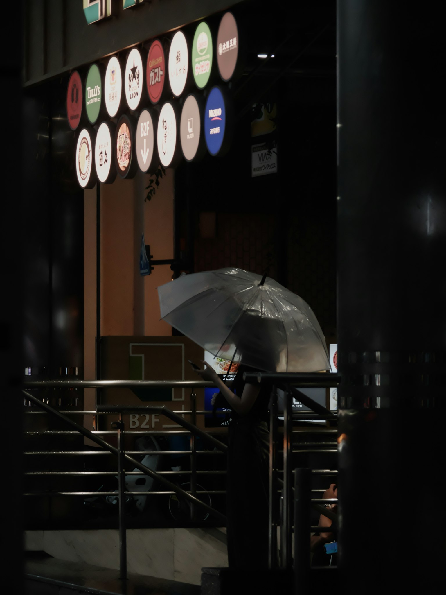 Une personne tenant un parapluie se tient sur des escaliers la nuit Divers panneaux colorés sont visibles en arrière-plan