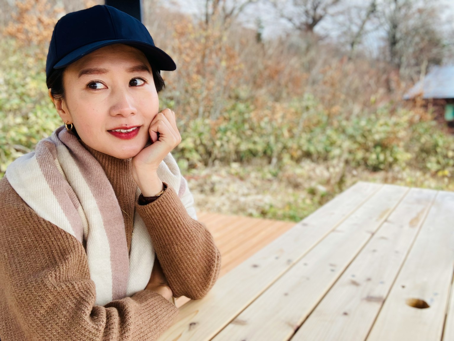 캐주얼한 복장의 여성이 모자 쓰고 나무 테이블에 앉아 미소 짓고 있음