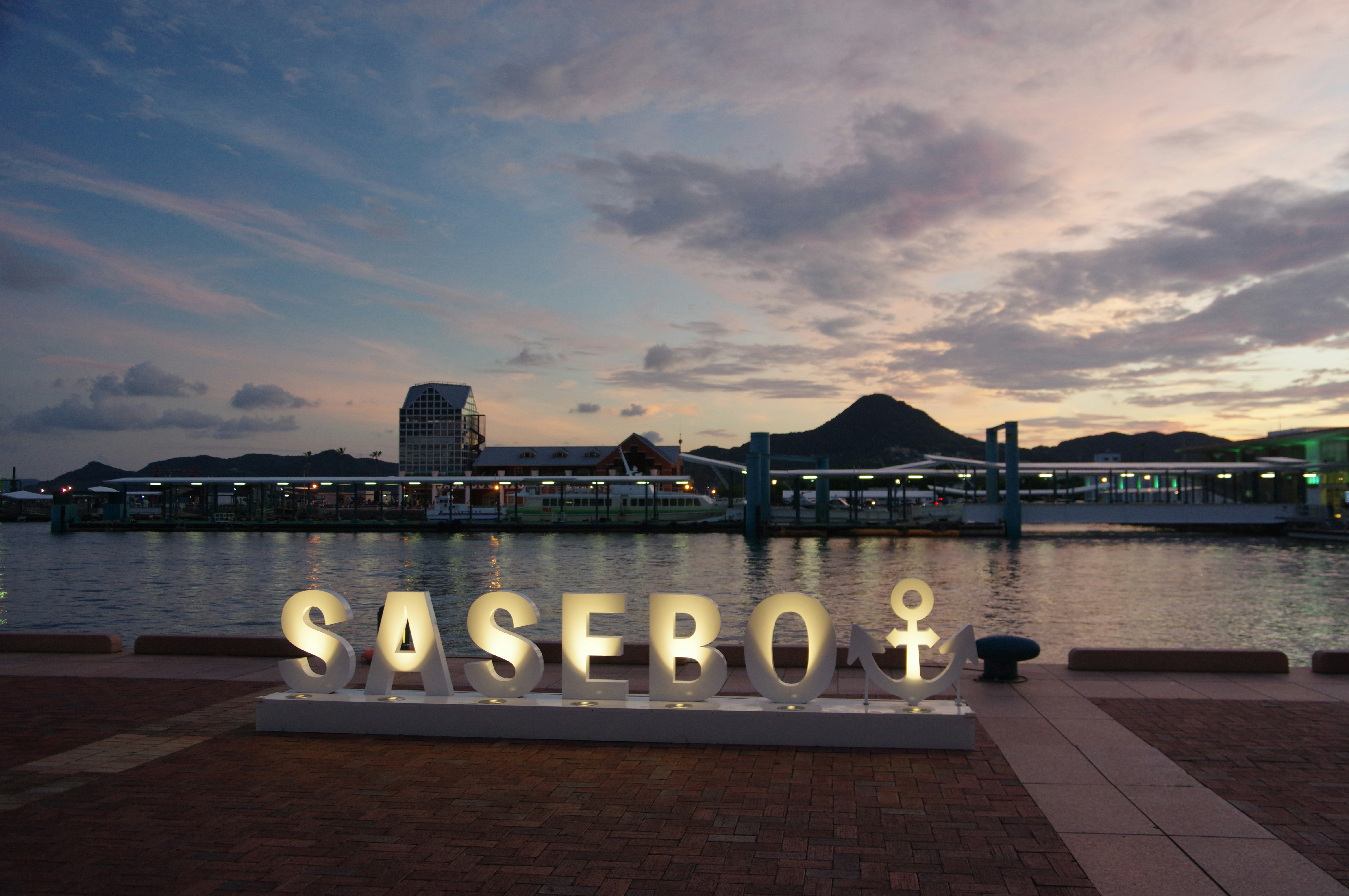 papan bertuliskan “SASEBO” di tepi perairan saat senja