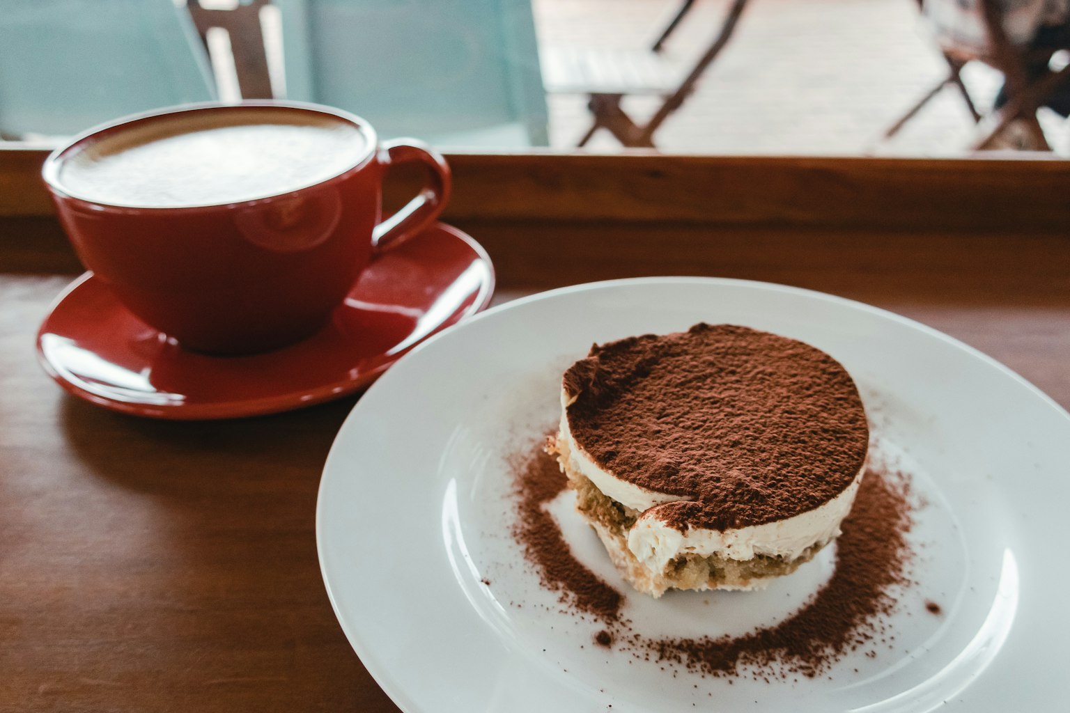 Cốc cà phê đỏ bên cạnh đĩa tiramisu trên khay gỗ trong không gian quán cà phê
