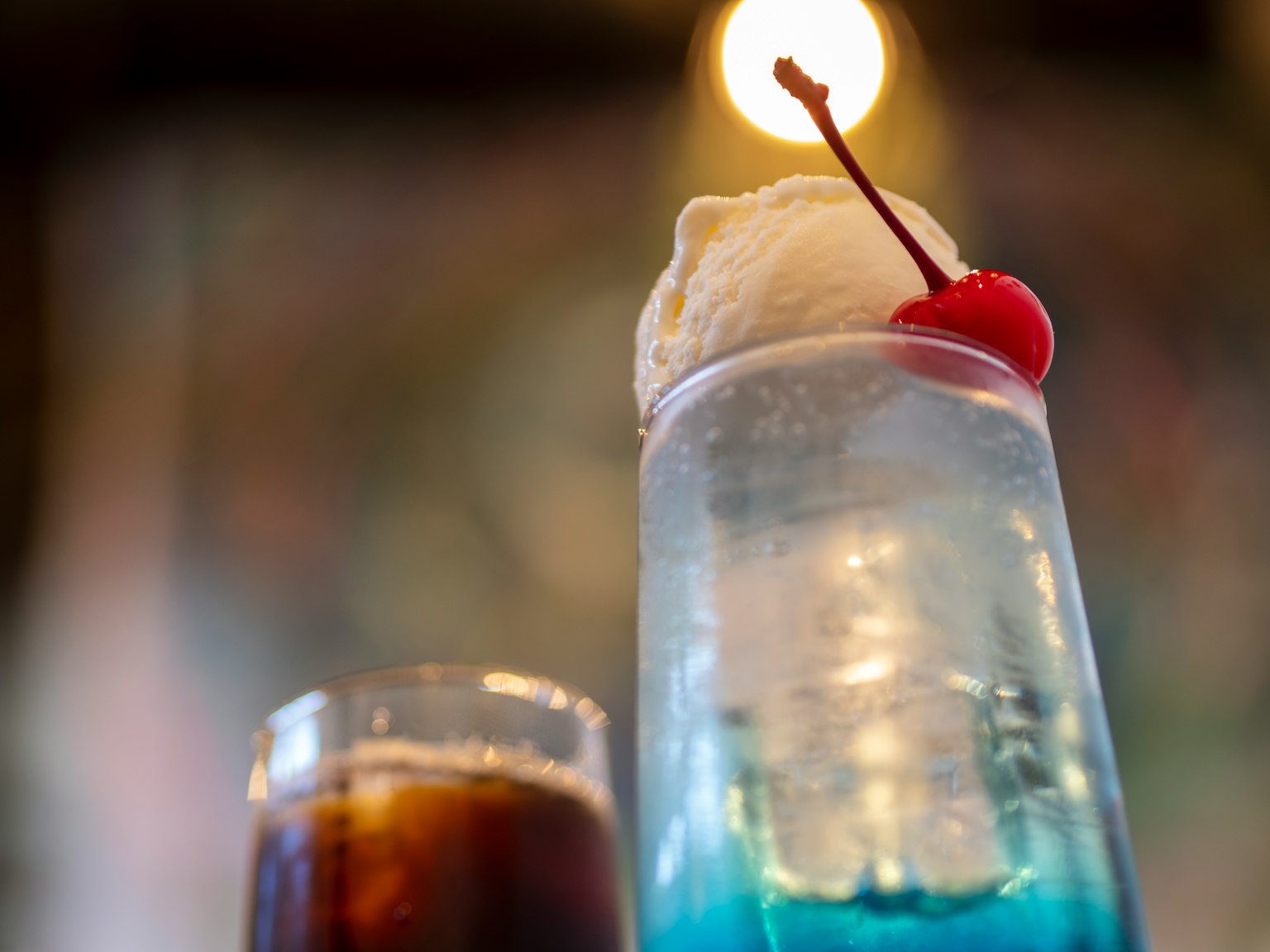 Một ly cocktail với đồ uống màu xanh và kem phủ một quả cherry bên cạnh một ly cola