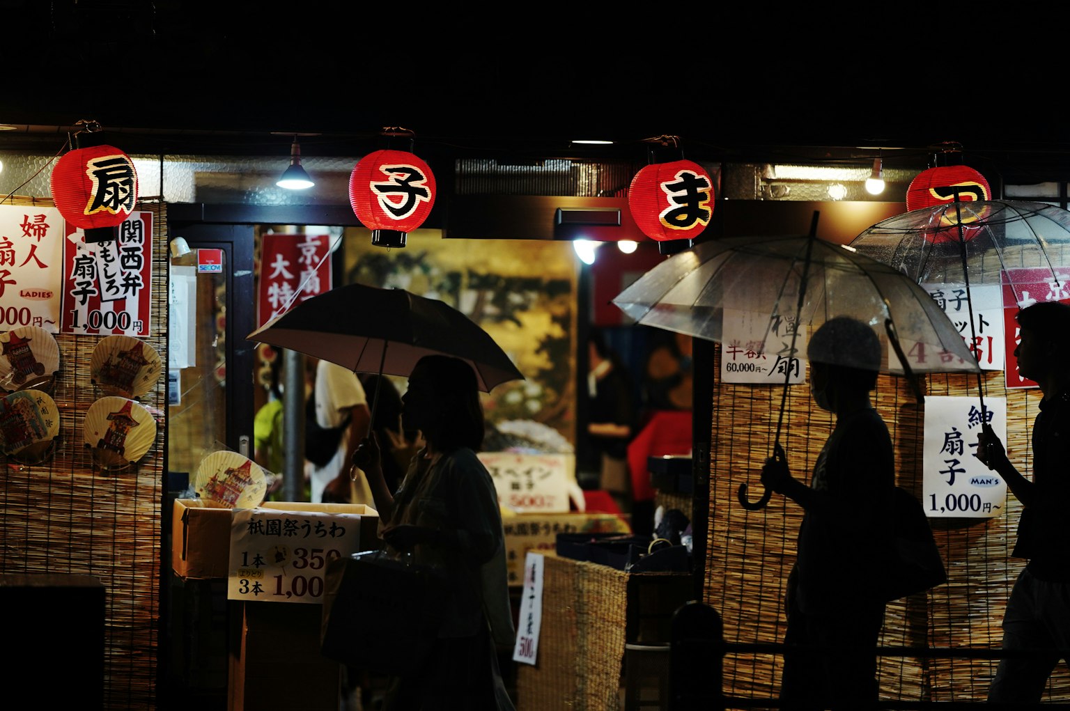 Silhouettes of people with umbrellas at a night market with glowing lanterns