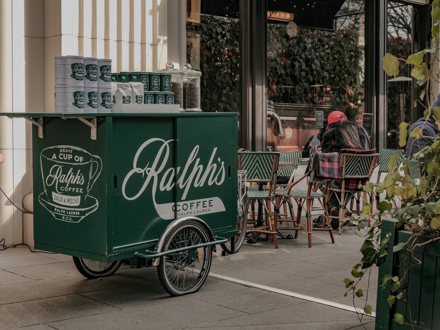 緑色のコーヒーカートに「Ralph's Coffee」のロゴがあり、屋外のカフェのテーブルが見える