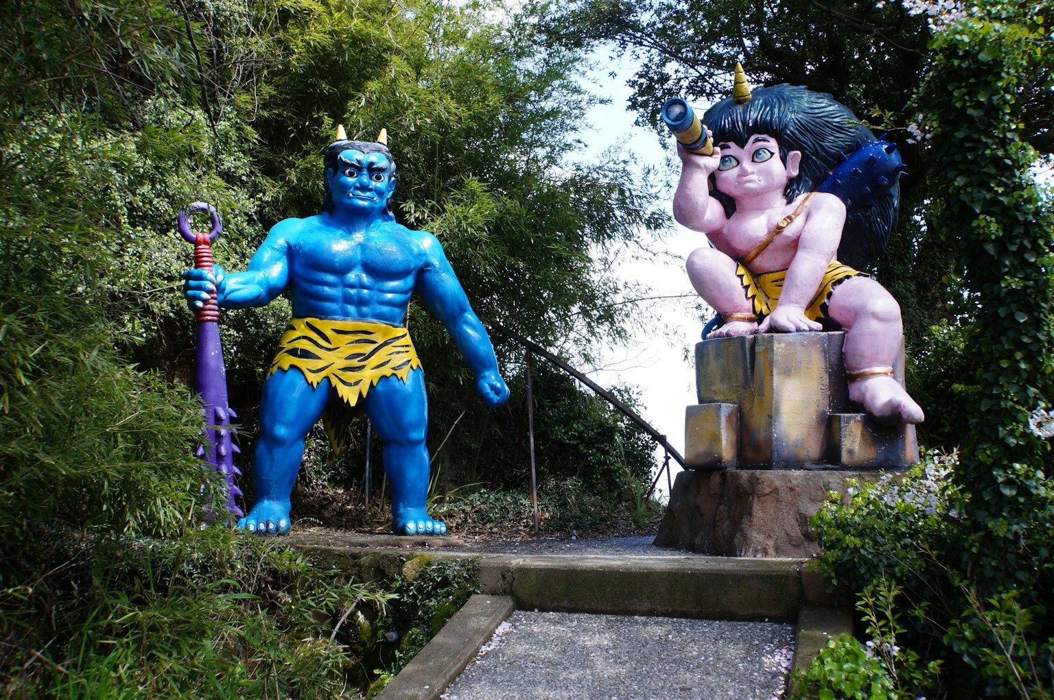 Imagen de un gigante azul y una estatua de deidad rosa en un entorno exterior exuberante