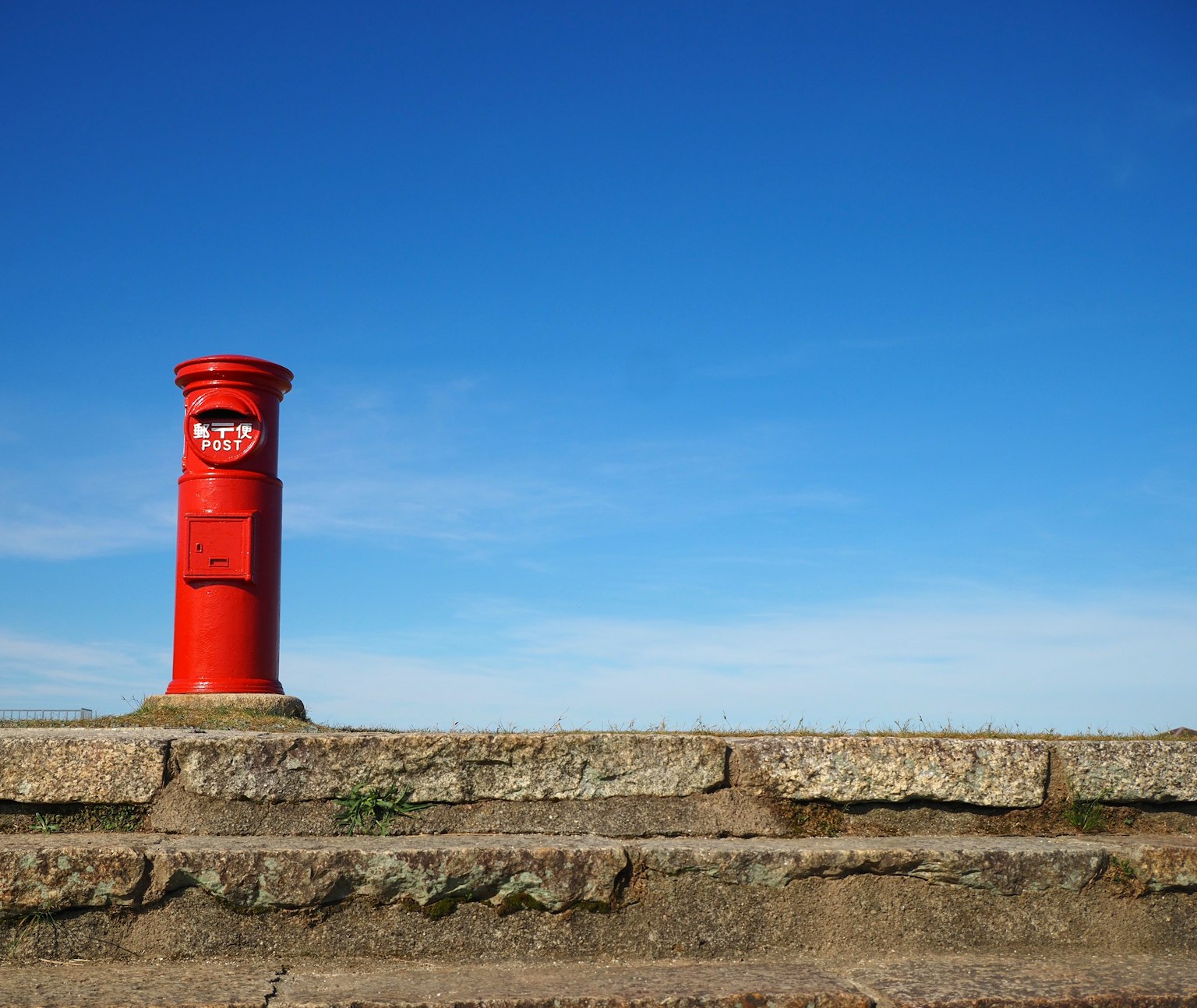 A red post box standing on stone steps against a blue sky background