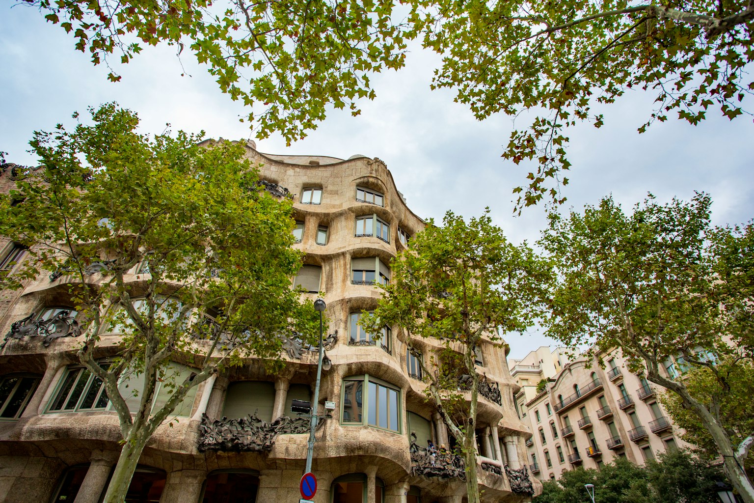 Exterior view of Casa Mila in Barcelona surrounded by green trees and a cloudy sky