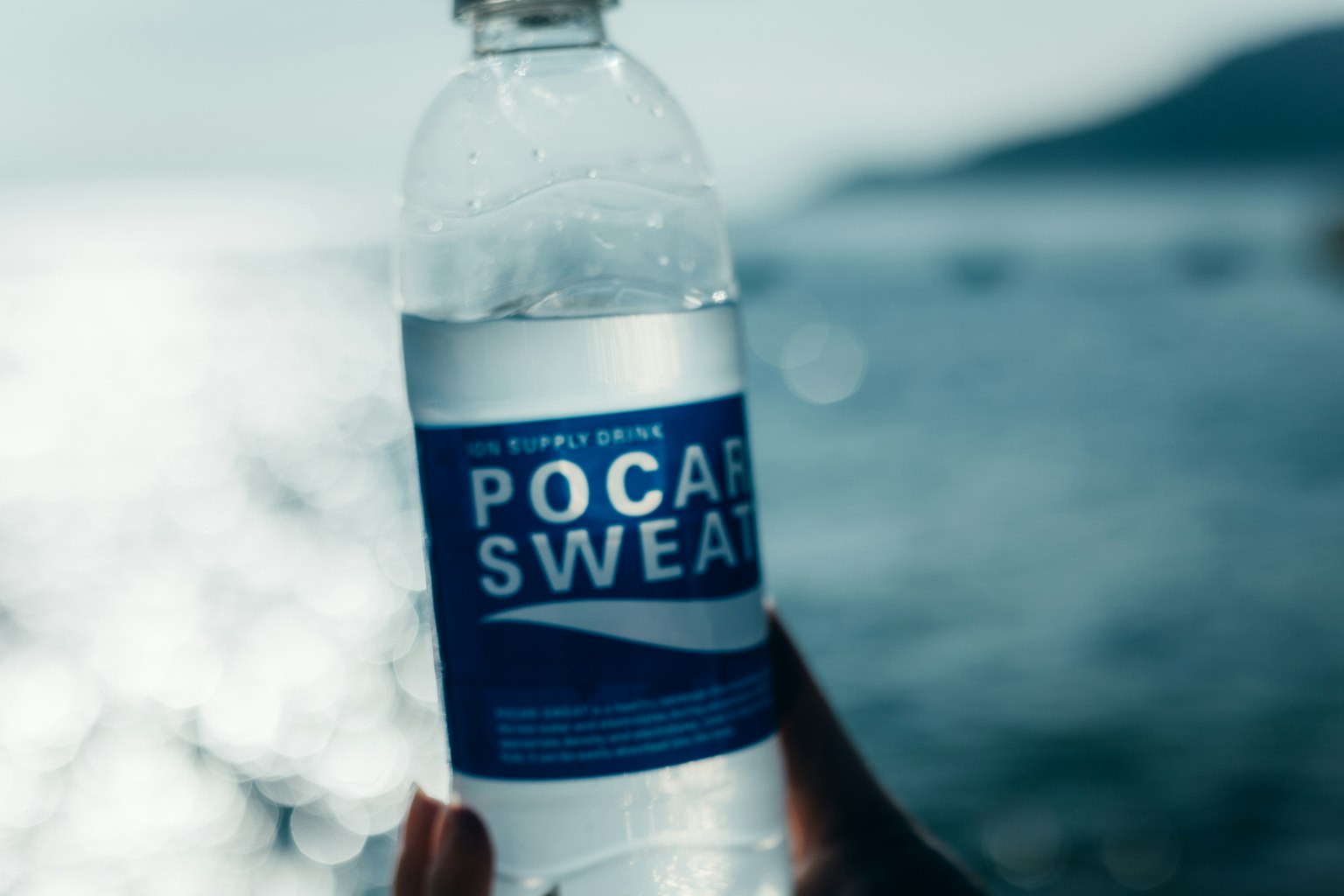 Hand, die eine Pocari Sweat-Flasche hält, mit verschwommenem Ozeanhintergrund