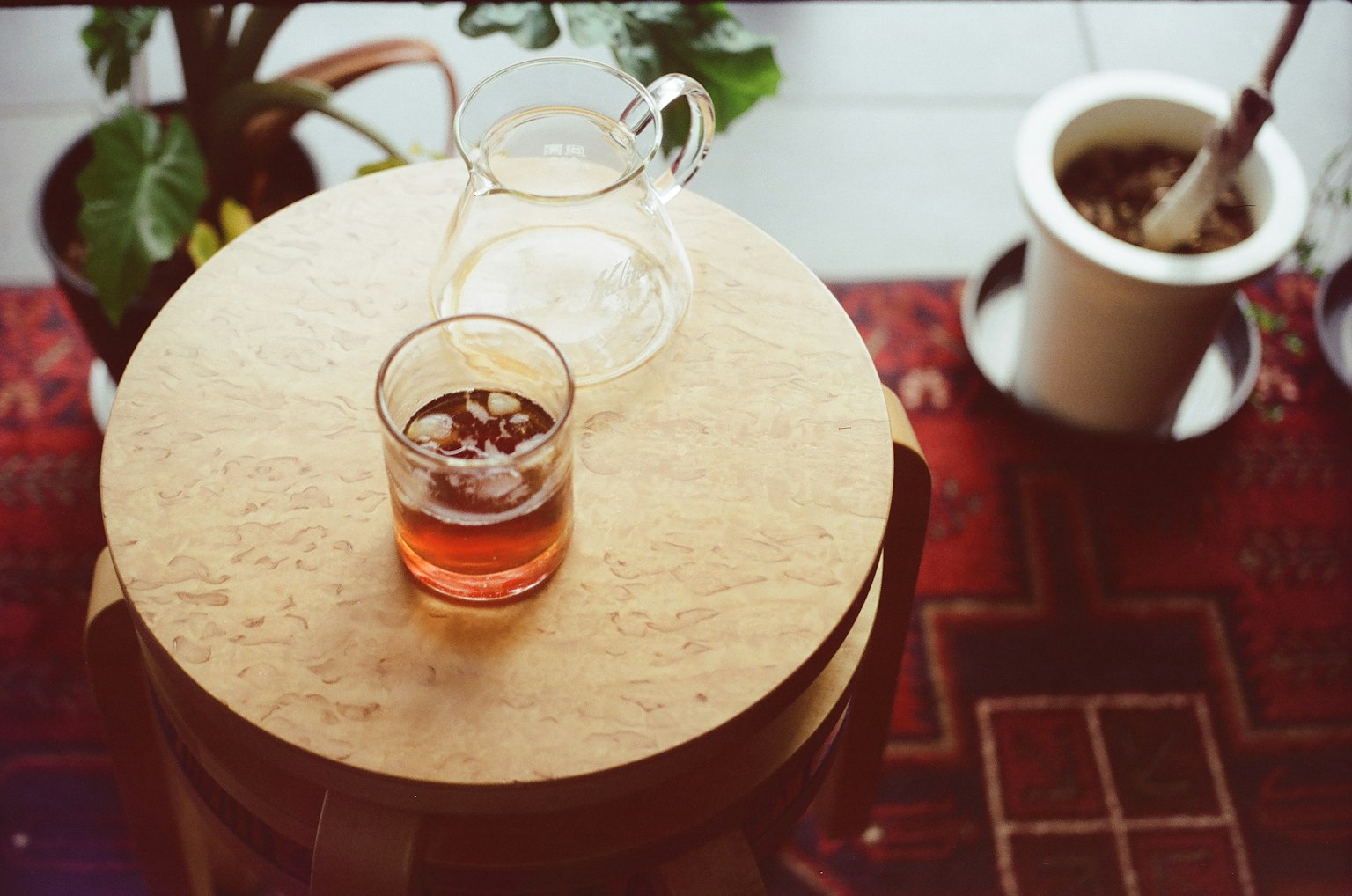 Segelas minuman es dan sebuah pitcher di atas meja kayu dengan tanaman dan mangkuk di latar belakang
