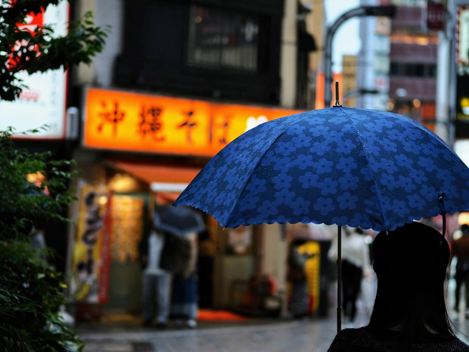 在雨中撐著藍傘的人影，背景有明亮的招牌