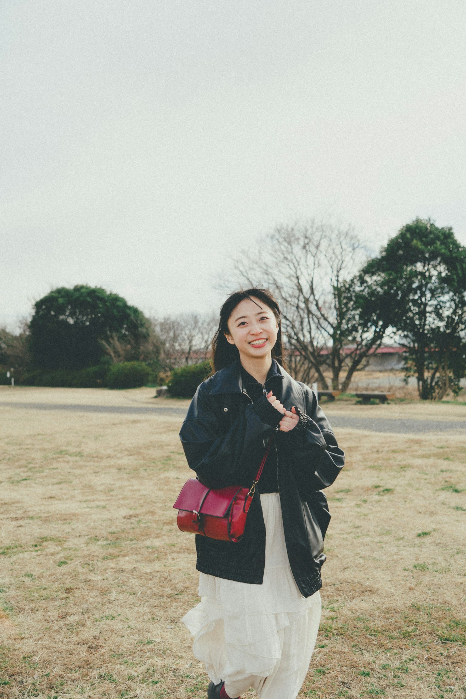 一名女性在公园里微笑着走着，穿着黑色夹克和白色裙子，手提红色包