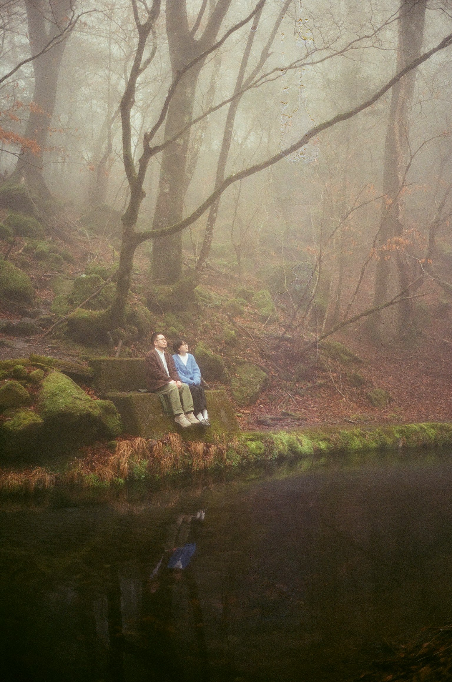 Two individuals sitting by a tranquil water body in a foggy forest