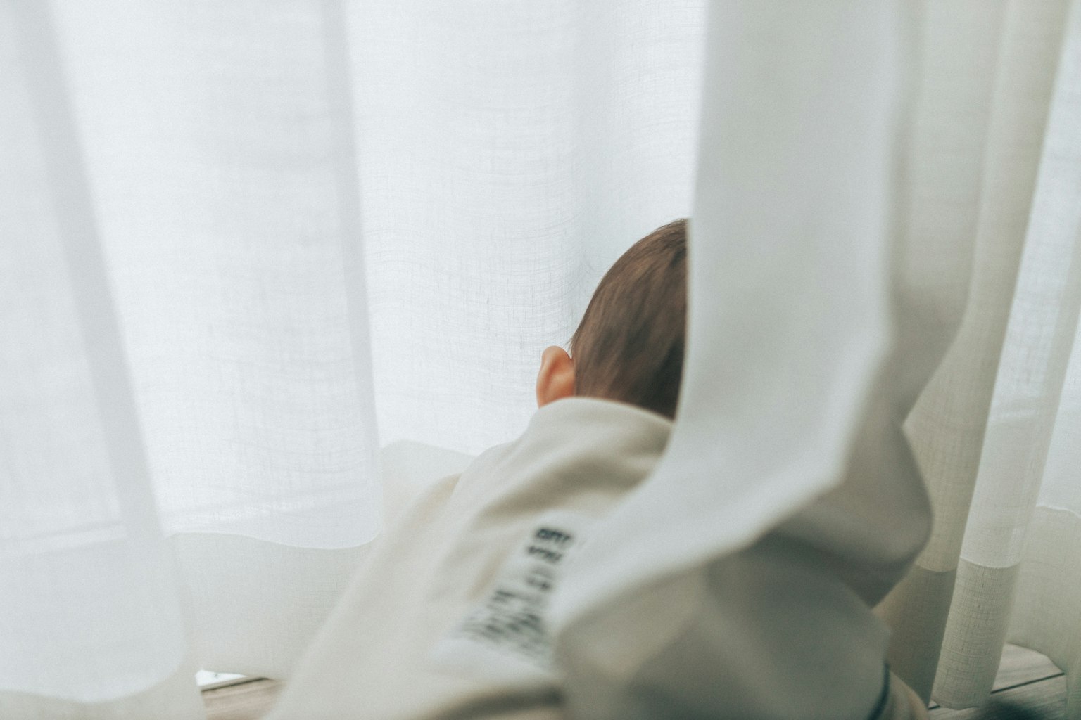 Testa di bambino che sbircia dietro tende bianche vicino a una finestra