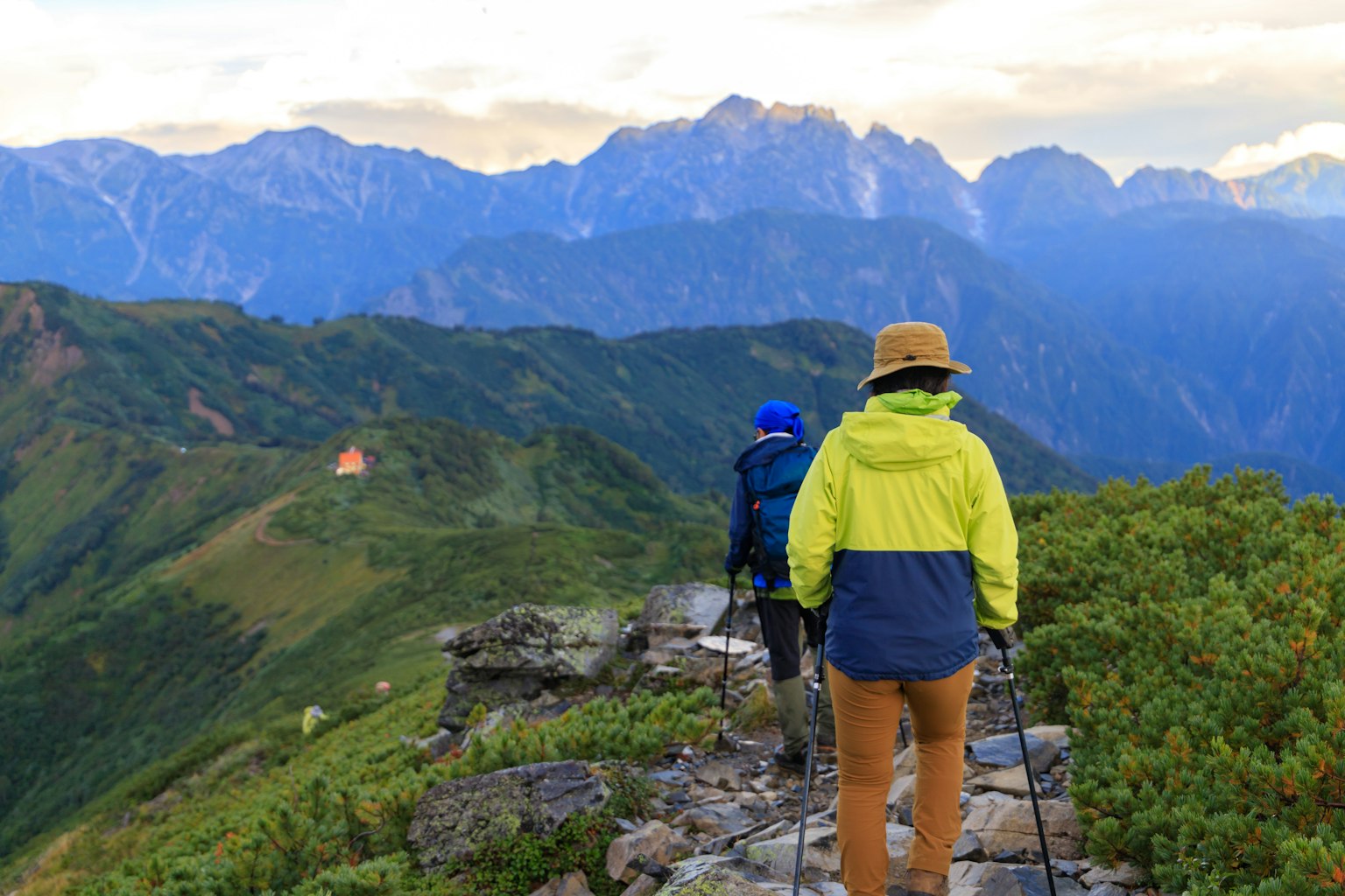 Dua pendaki berjalan di jalur gunung dengan padang rumput hijau dan gunung di kejauhan