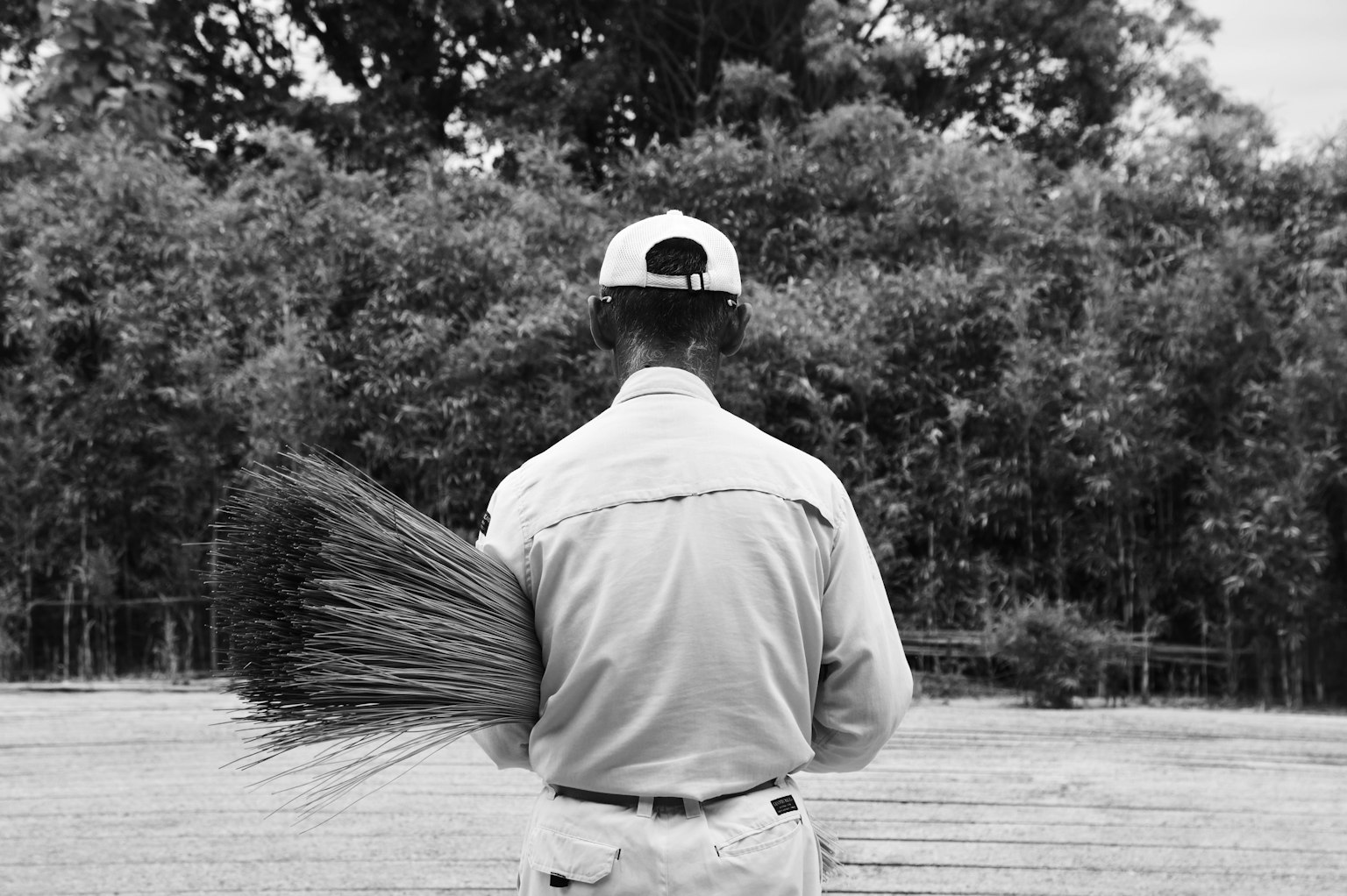 Un hombre sosteniendo una escoba visto desde atrás en una imagen en blanco y negro