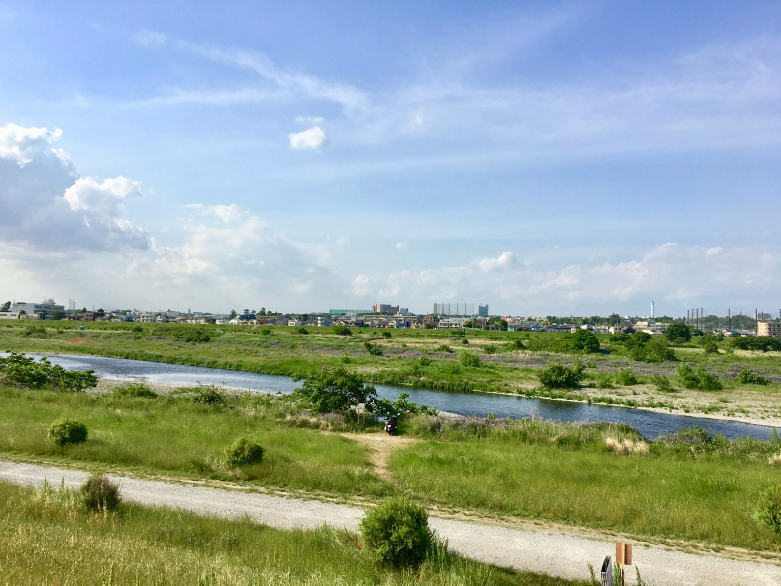 Ladang hijau yang luas di bawah langit biru dengan sungai yang mengalir dan bangunan di kejauhan