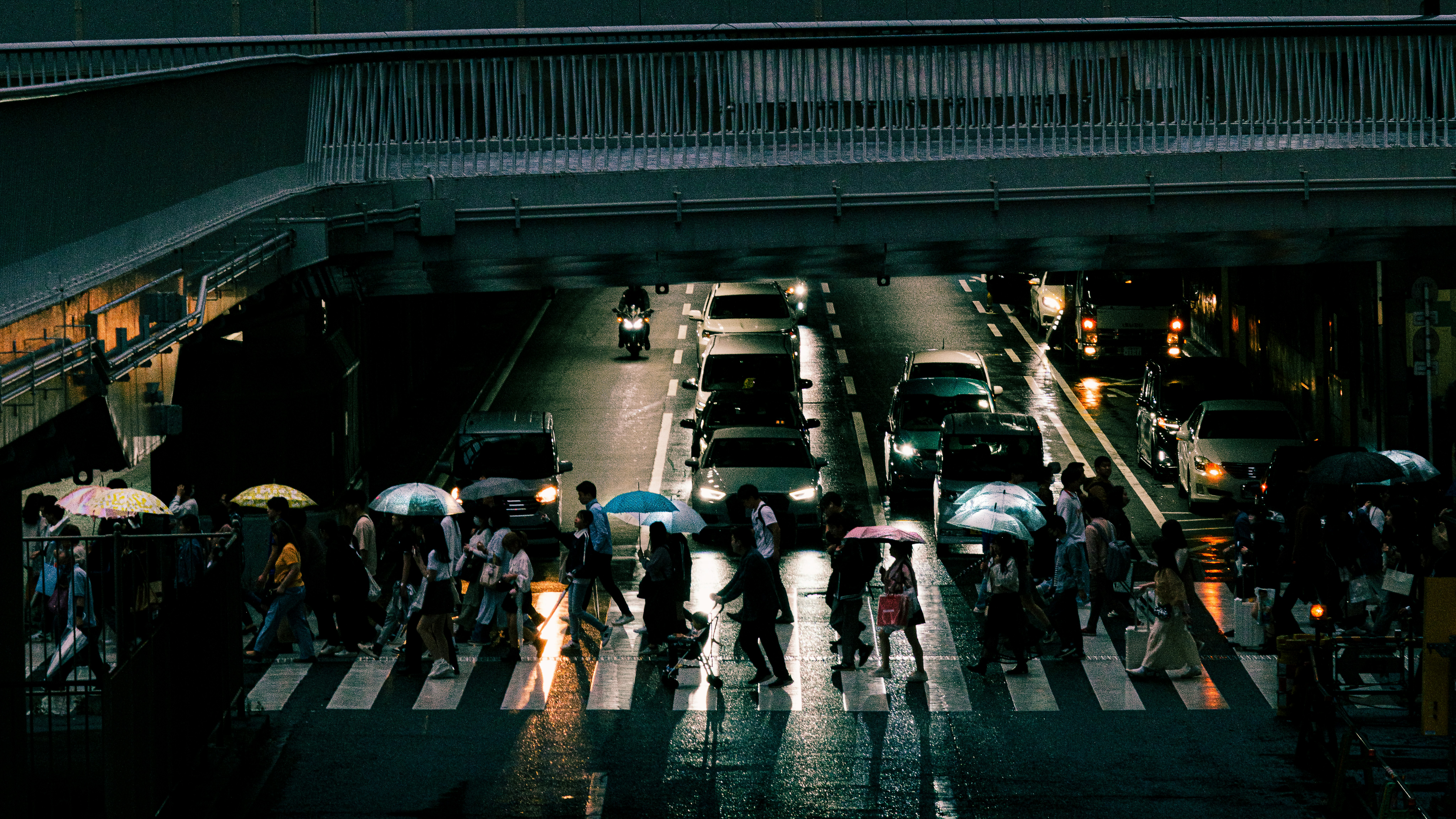 高架下の雨の駅前で傘をさす人々の行き交い