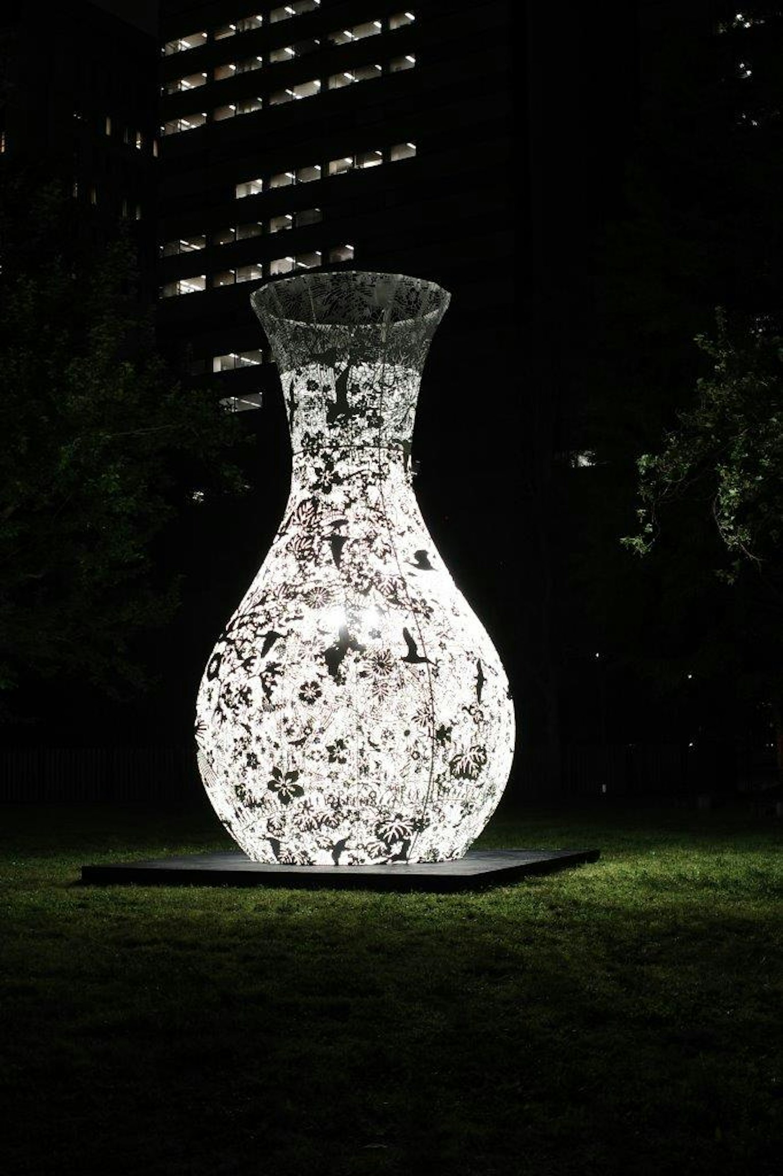 Grande scultura di vaso illuminato di notte circondata da erba verde e grattacieli
