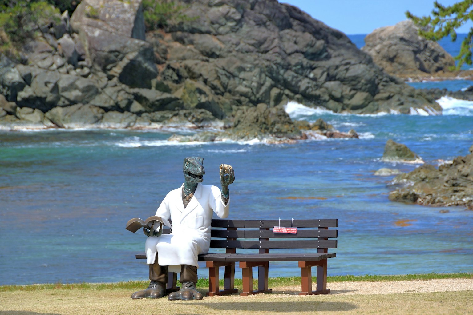 Una estatua con un abrigo blanco sentada en un banco junto a la playa con una mano levantada