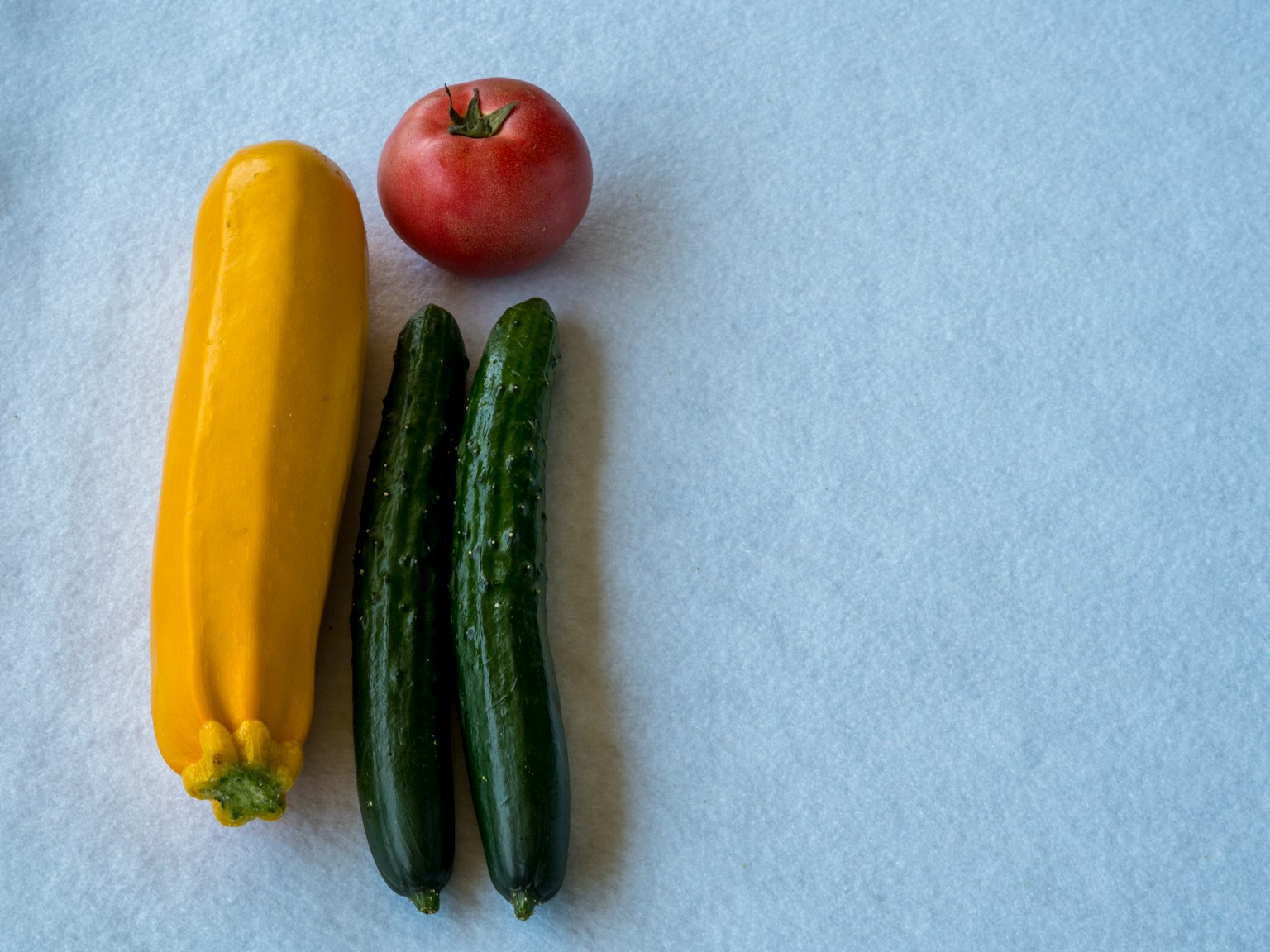 Sebuah zucchini kuning, satu tomat merah, dan dua mentimun hijau disusun di atas latar belakang putih