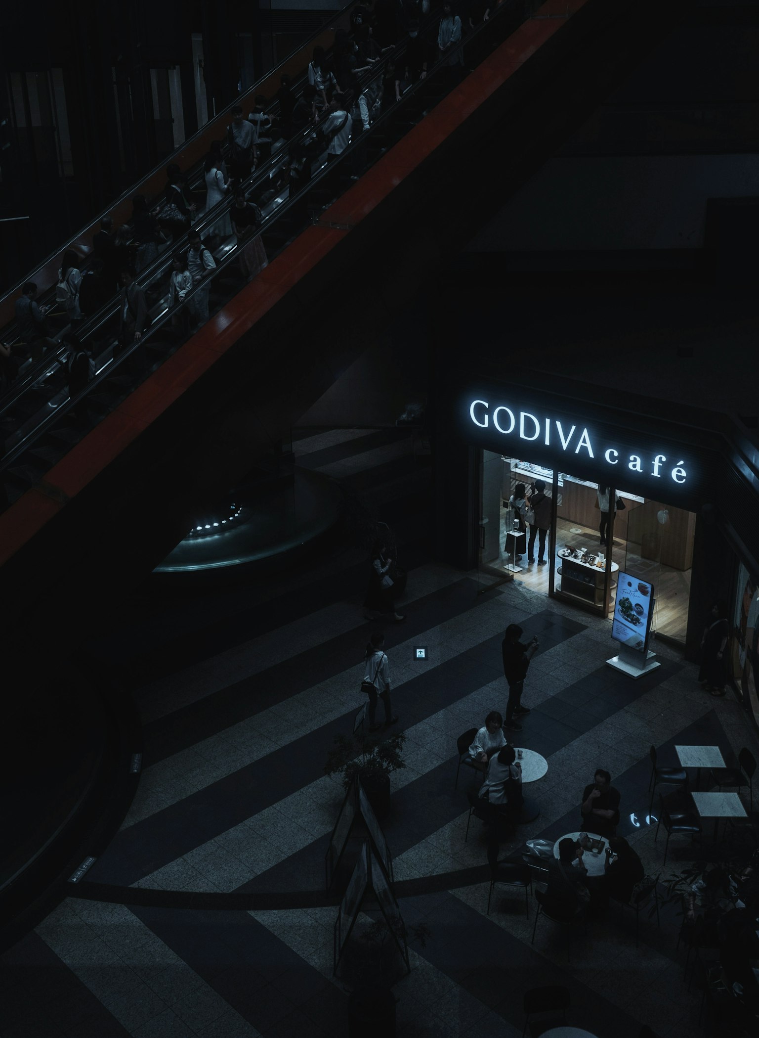 Papan kafe GODIVA di ruang yang redup dengan orang-orang yang berjalan