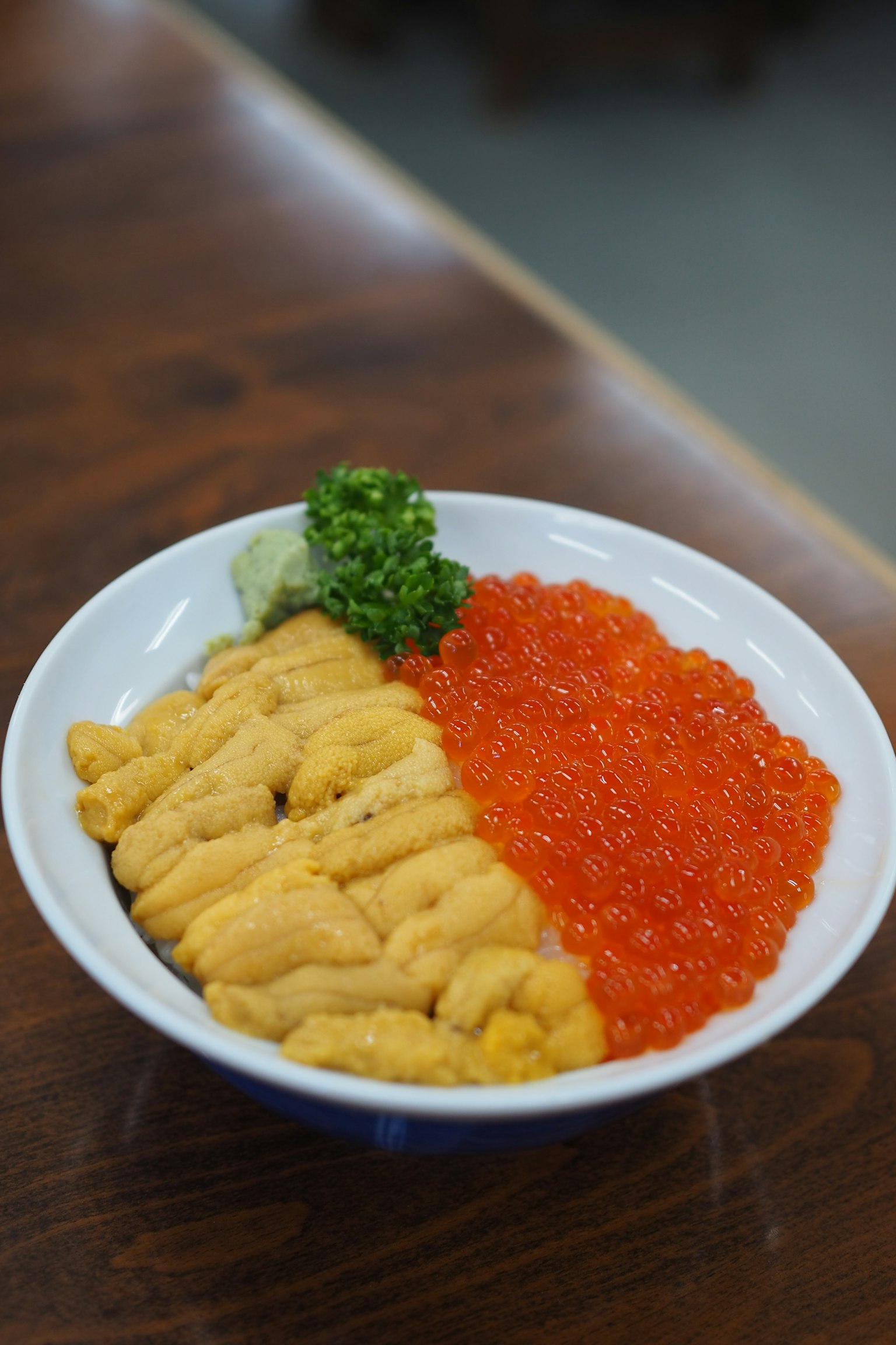 Mangkuk berisi uni dan telur salmon dengan wasabi hijau dan daun dalam piring keramik putih