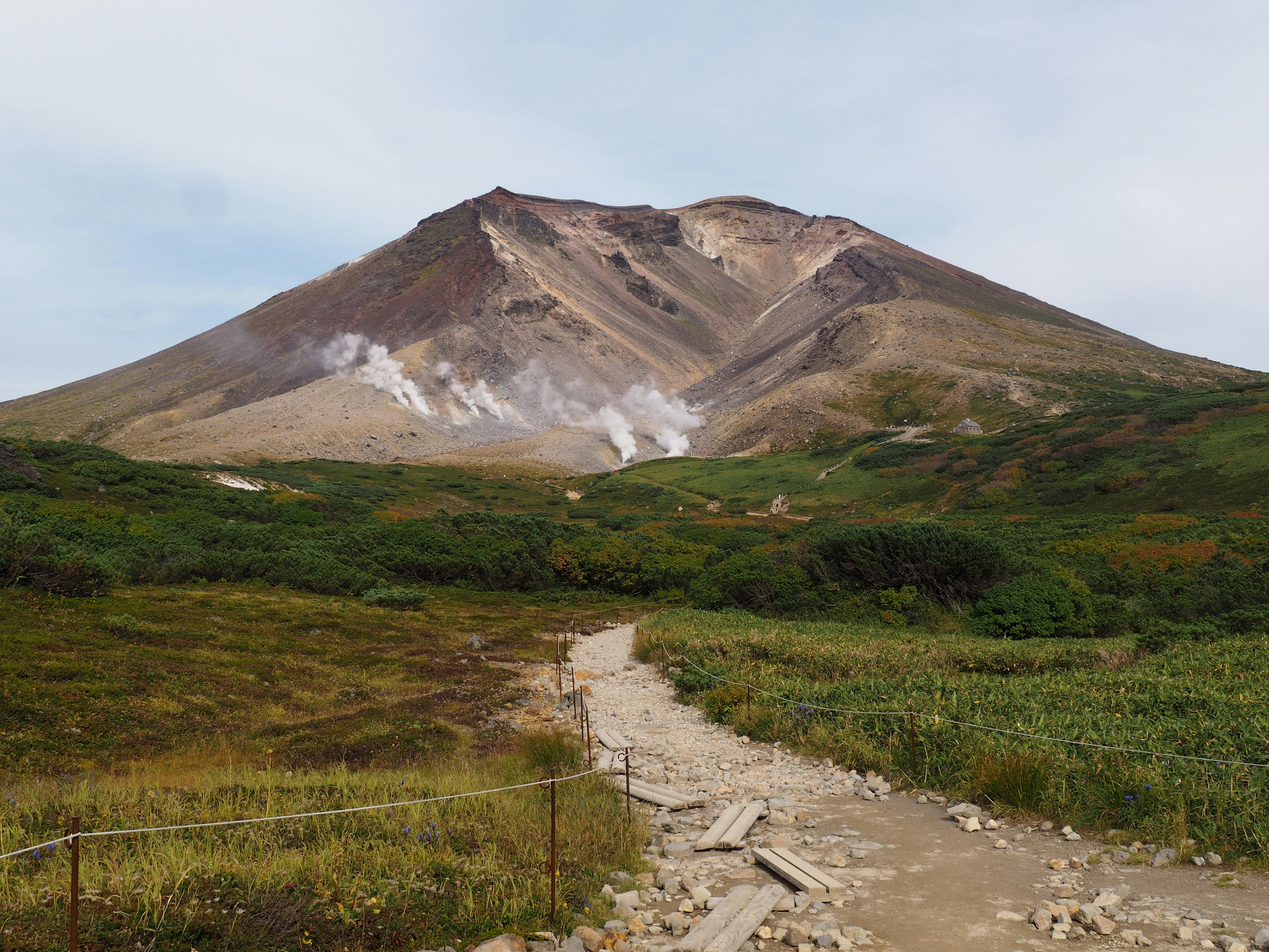 火山の噴気と荒れた山肌を望む登山道のある自然風景