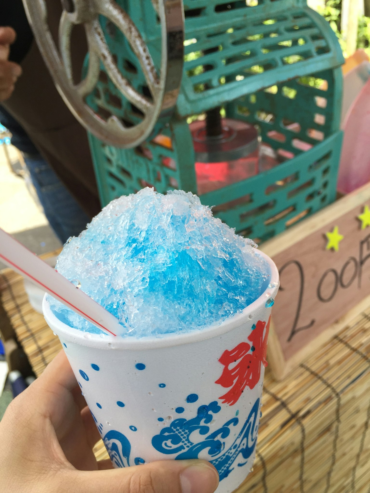 Hand holding a cup of shaved ice topped with blue syrup vintage ice shaver and sign in the background