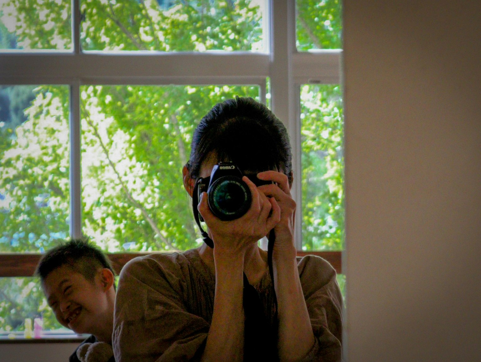Self-portrait of a person holding a camera with green trees visible through the window