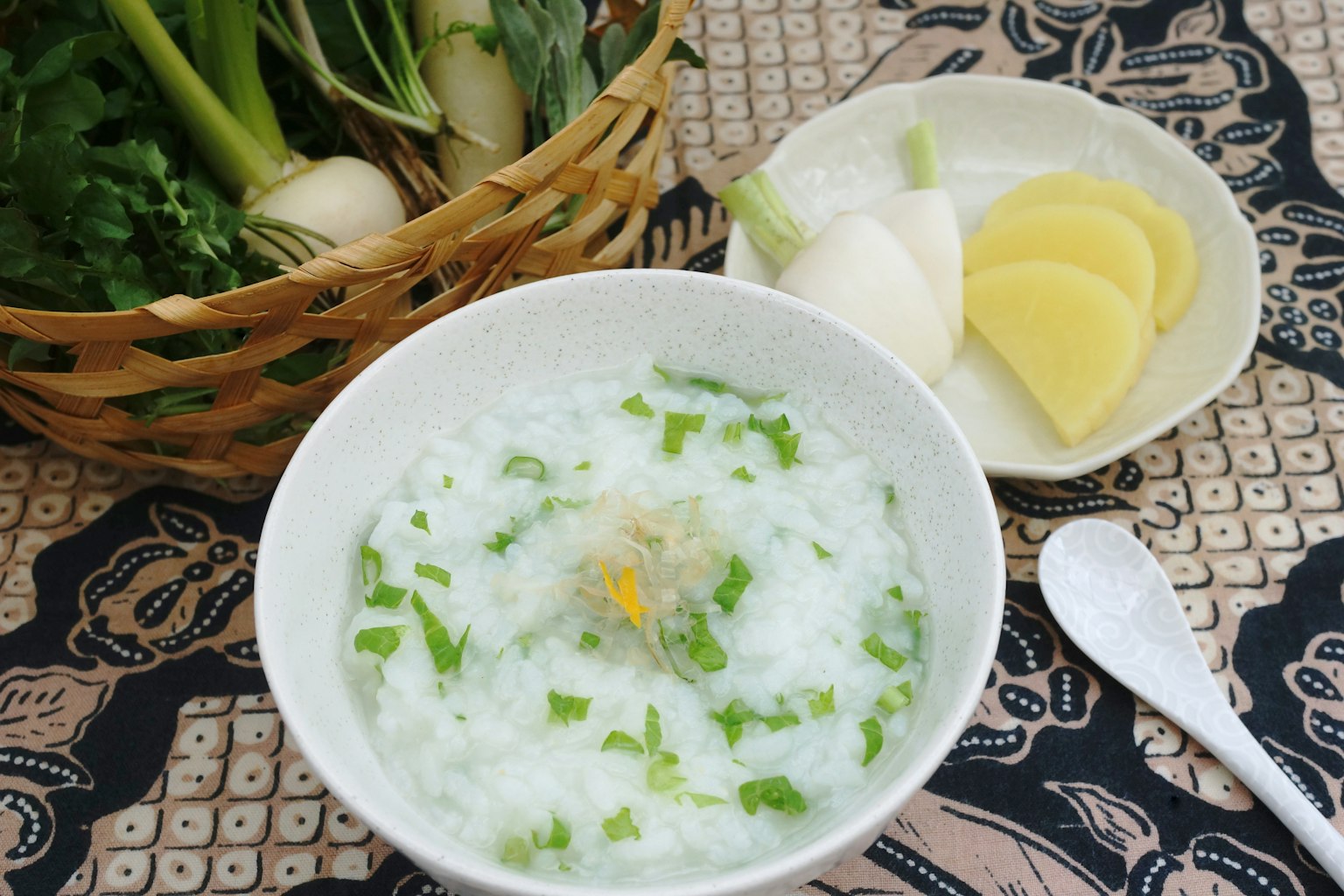 Ciotola di porridge di riso bianco guarnita con cipollotti e erbe accanto a fette di ravanello e patata