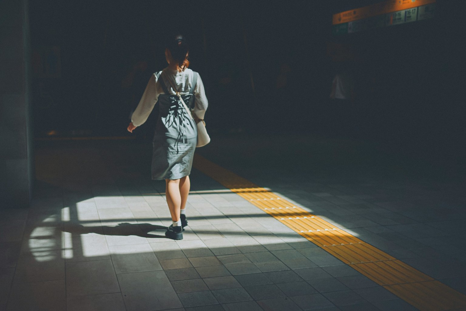 薄暗い駅の中を歩く女性の後ろ姿 明るい黄色のラインが床に映る