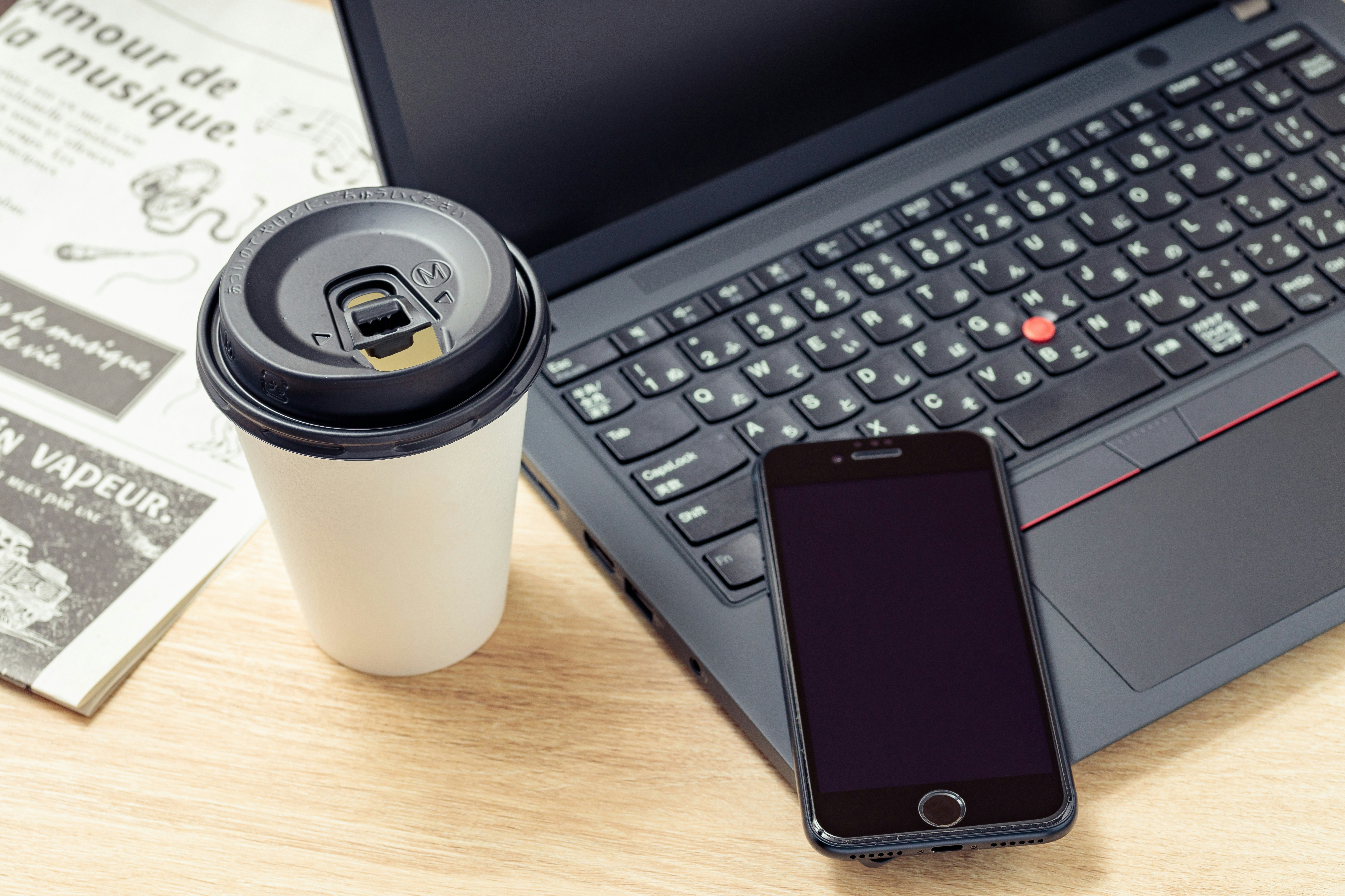 ノートパソコン、コーヒーカップ、スマートフォン、新聞が並ぶデスクの上のシーン