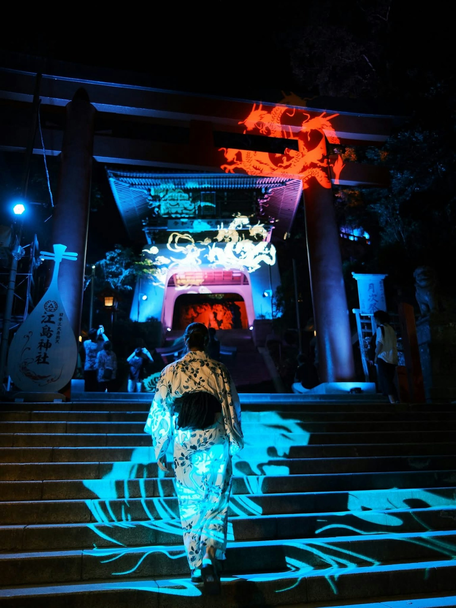 Một người phụ nữ trong trang phục kimono đang đi lên bậc thang của một ngôi đền vào ban đêm với những ánh sáng màu sắc