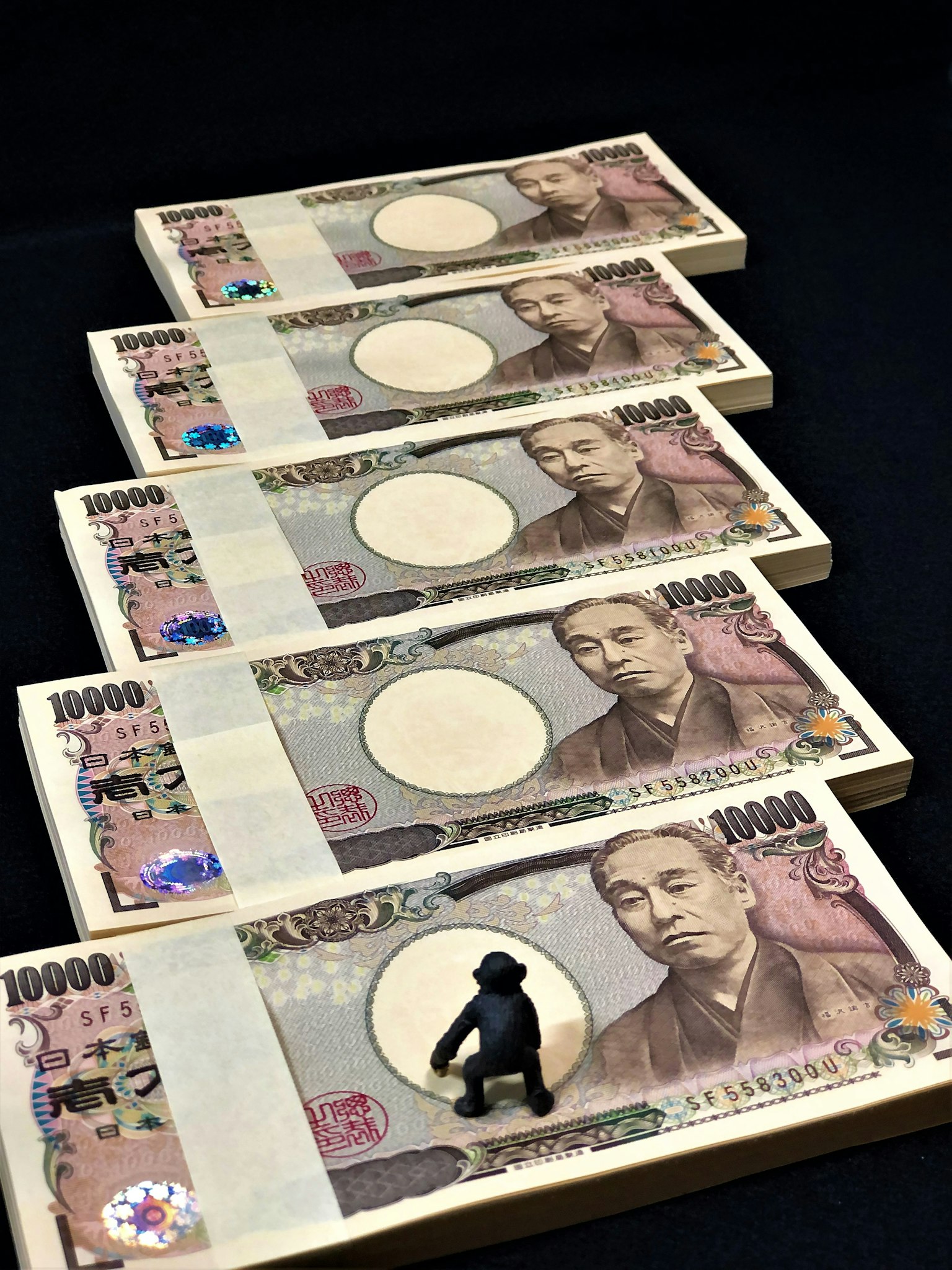 五張日本10000日圓鈔票排列在黑色背景上，上面放著一個小猩猩模型