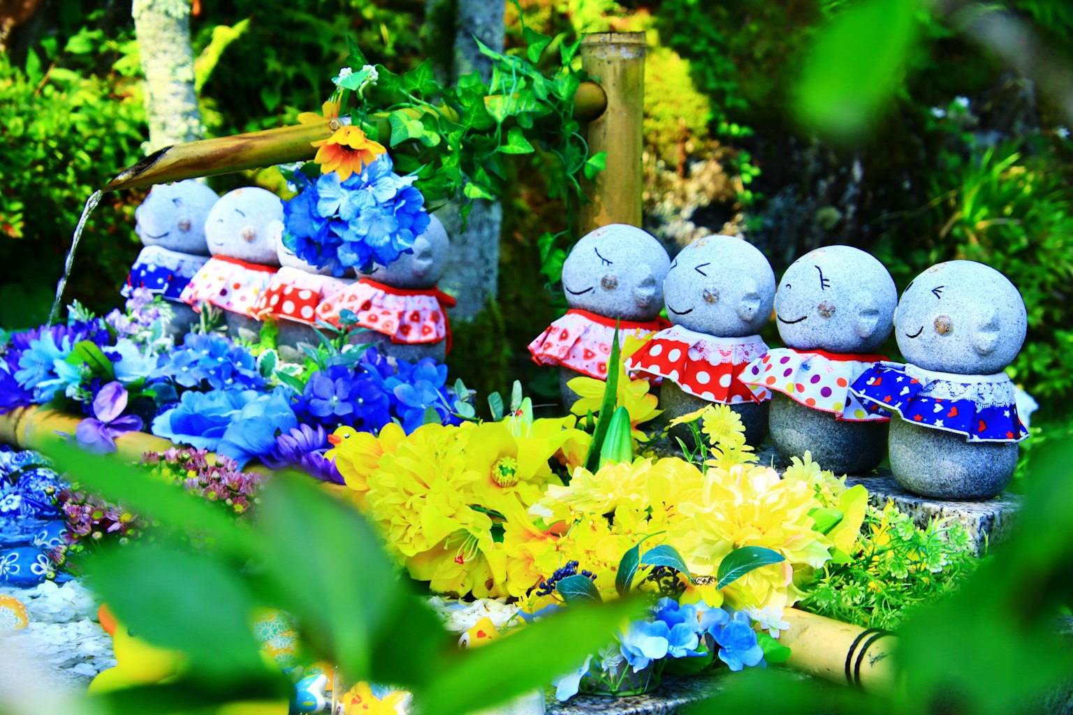 Petites figurines en pierre habillées de tissus colorés entourées de fleurs vibrantes créant une atmosphère joyeuse