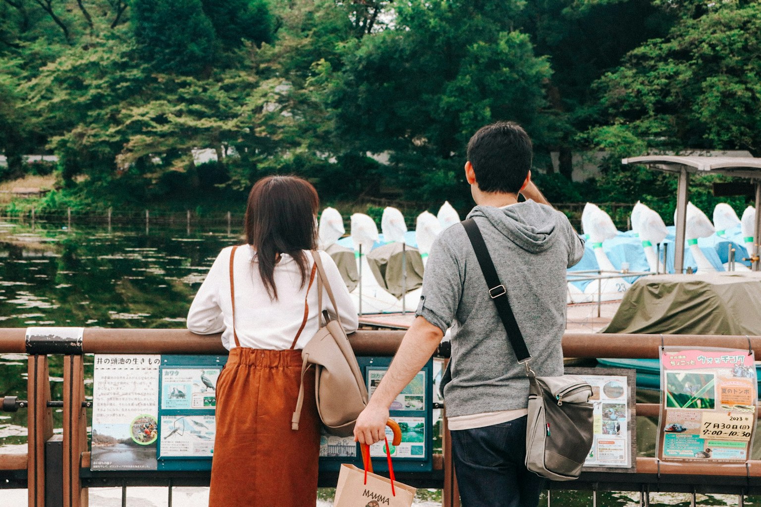 Cặp đôi đứng trước ao hồ trong công viên được bao quanh bởi cây cối và thuyền