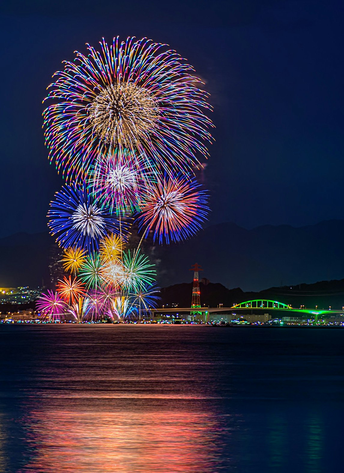 夜空に咲く花火と川沿いのライトアップの夜景