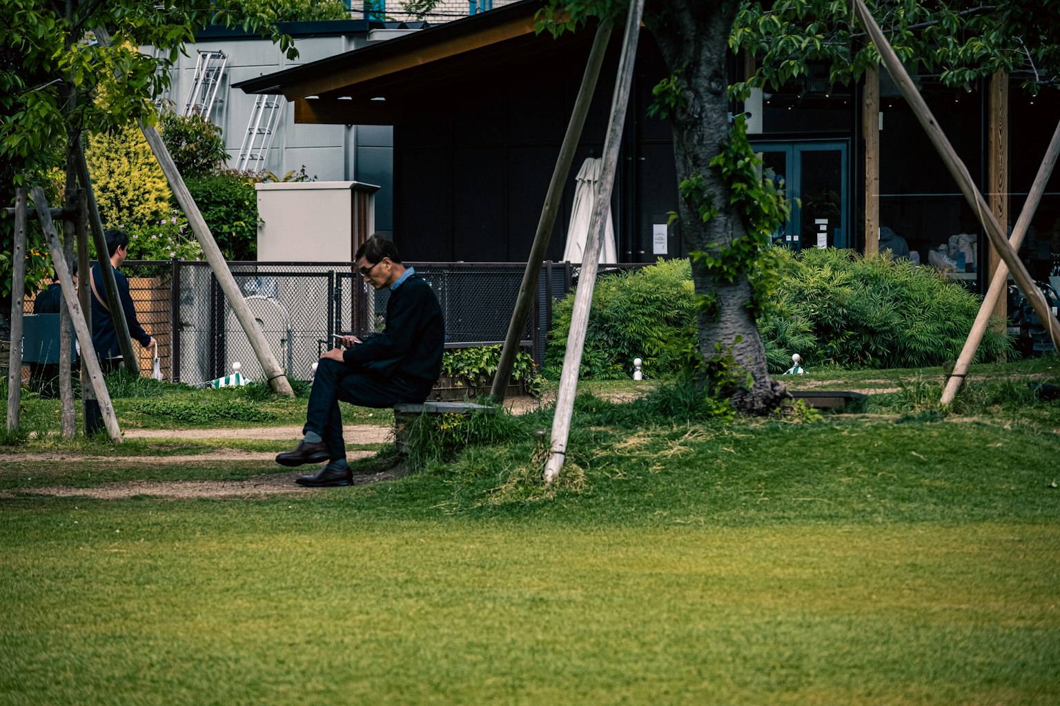公園のベンチに座る男性 スーツを着てスマートフォンを操作している 緑の芝生と木々が周囲にある