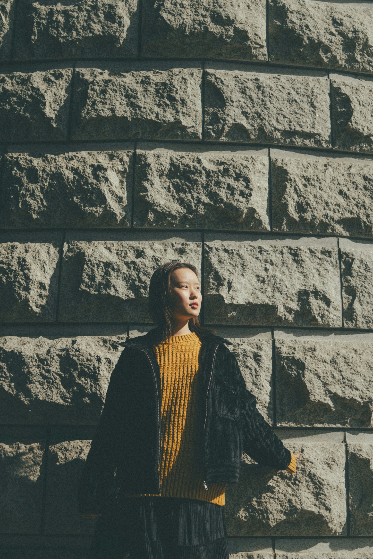 A woman standing in front of a stone wall wearing a bright yellow sweater and a black jacket