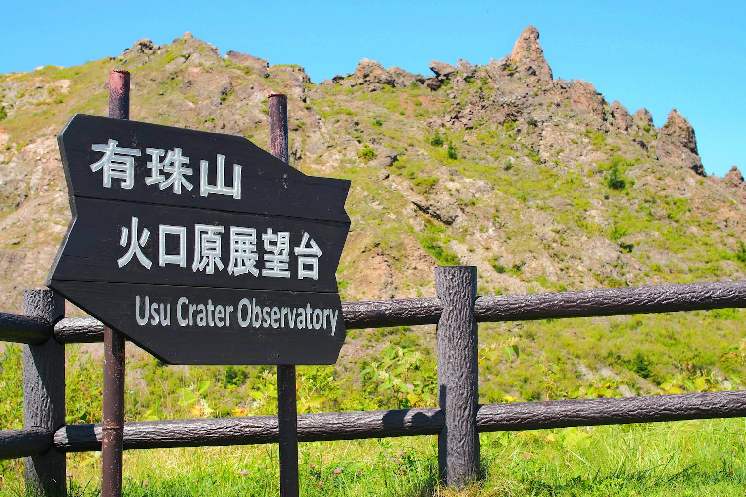 Cartel del Observatorio del Cráter de Usu con montañas verdes de fondo